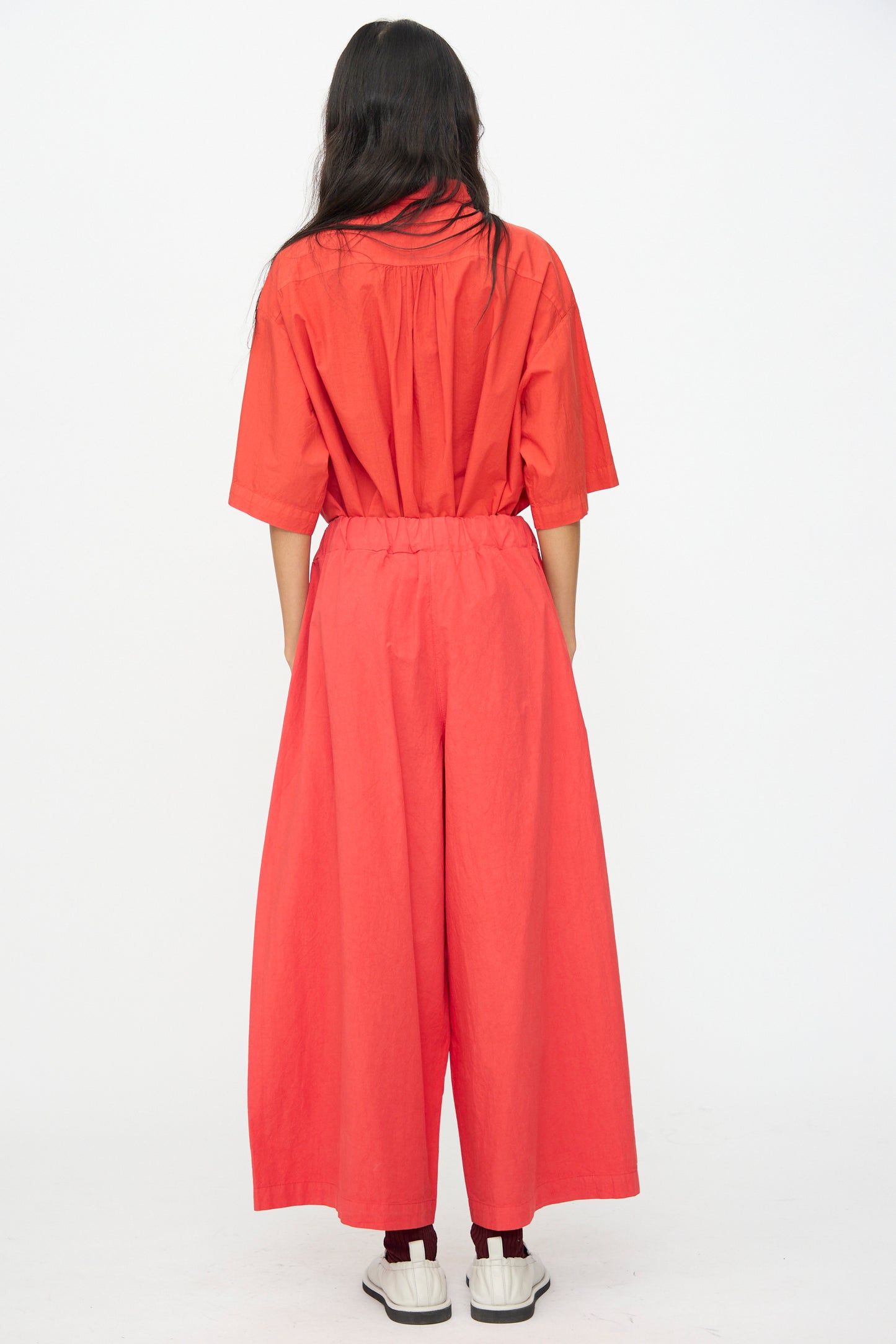 A person stands with their back to the camera, wearing the Labo.Art Cotton Sole Pant in Tango and white slip-on shoes against a plain white background.