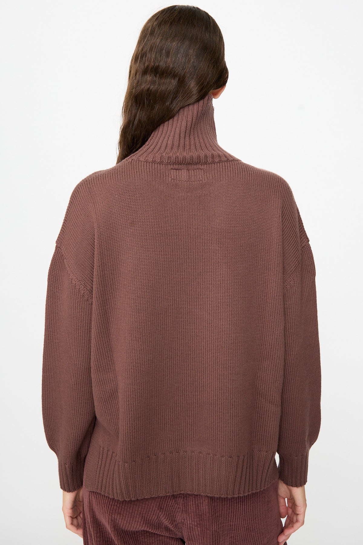 A person with long brown hair is photographed from the back against a plain white background, wearing the Labo.Art Fiore Sweater in Sasso—a loose, cashwool knitwear turtleneck.