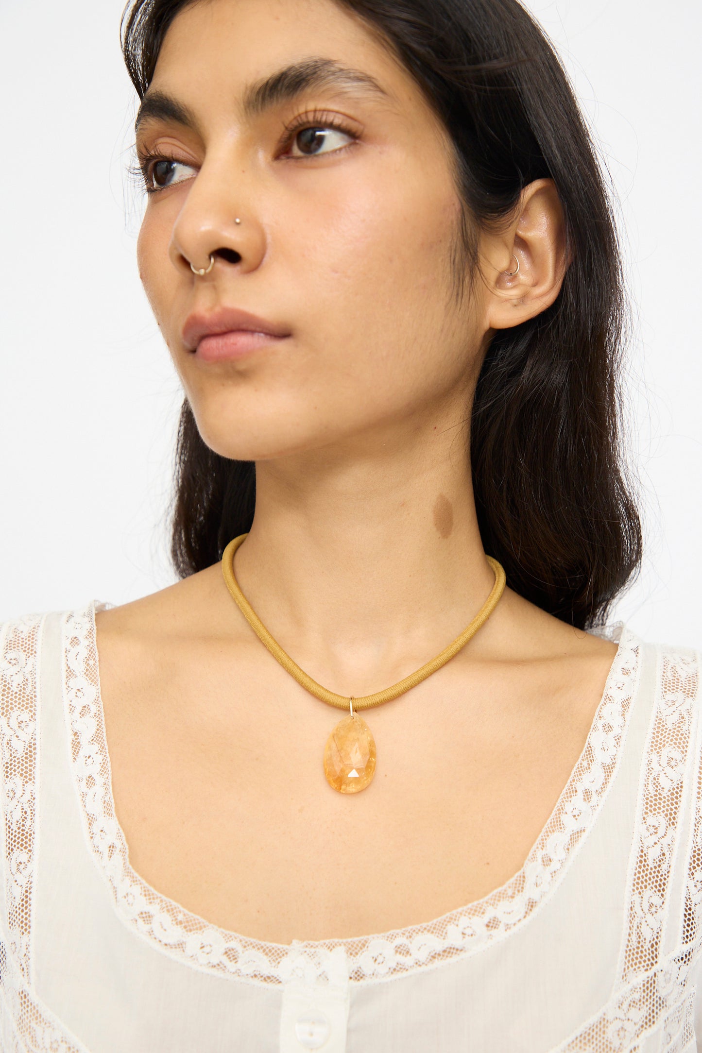 A person wearing a white lace-trimmed top and a gold necklace, The Drop Cord in Tan and Citrine by Laoli, featuring an elegant citrine pendant, shown from the shoulders up against a plain background.