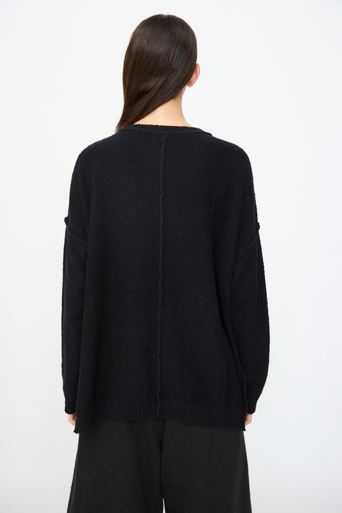 A person with long brown hair faces away, wearing the Lauren Manoogian Boucle Crewneck in Black and wide black pants, set against a plain light background.