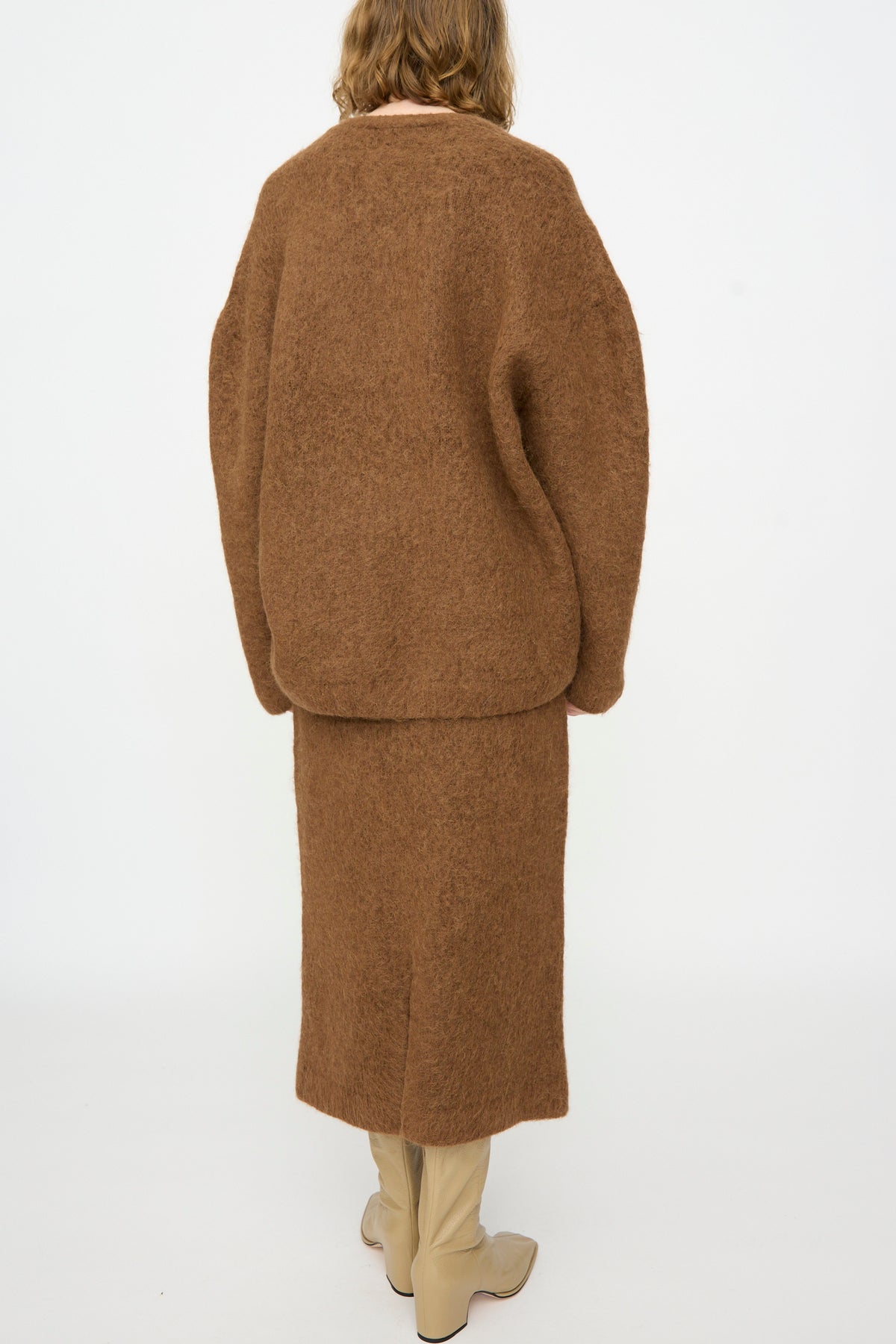 A person stands with their back to the camera, wearing the Lauren Manoogian Brushed V Neck in Rust — a loose, oversized Suri Alpaca sweater — paired with a matching brown skirt and cream knee-high boots, set against a plain white background.