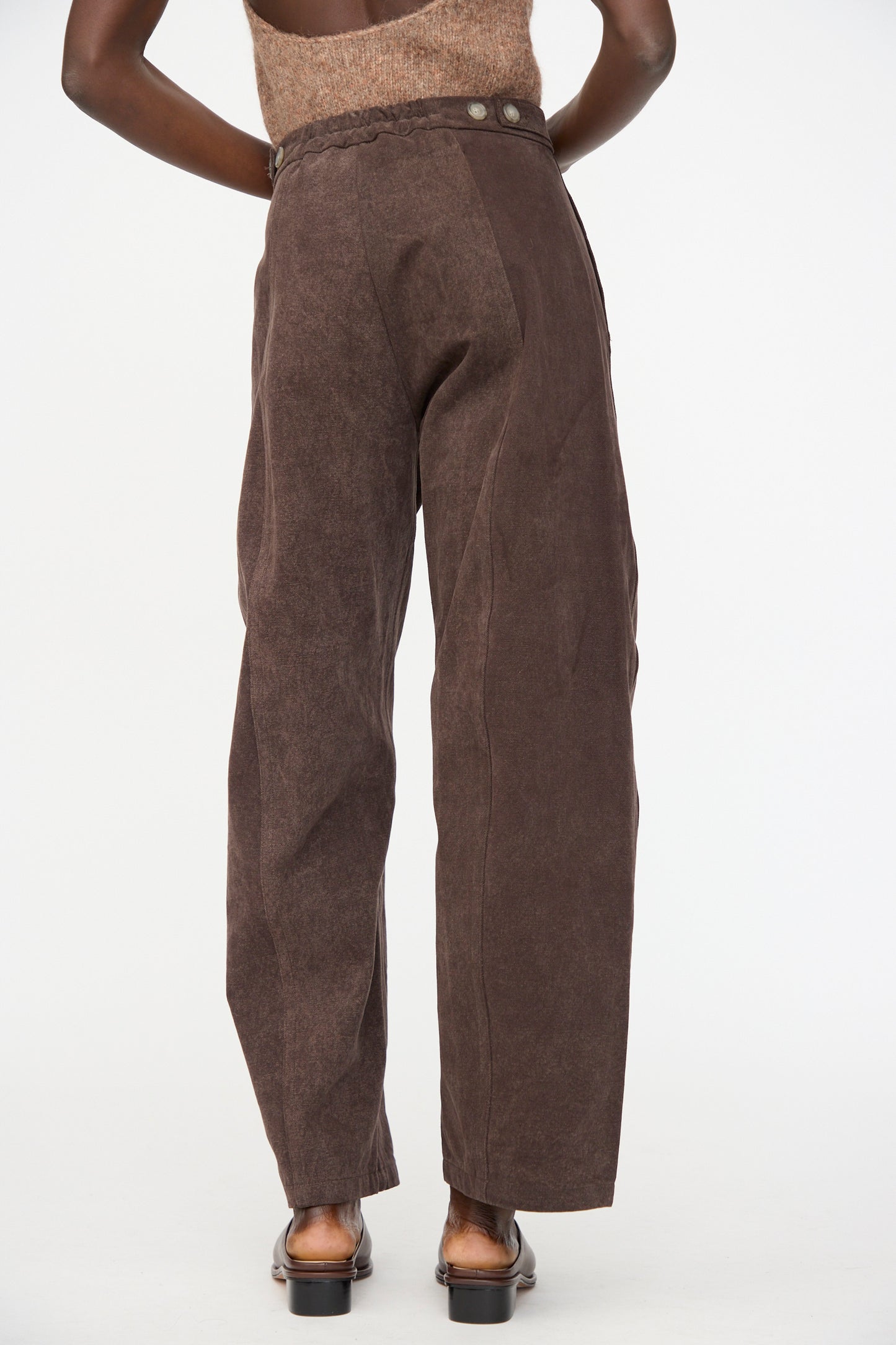 Someone stands with their back to the camera against a plain background, wearing Lauren Manoogian Canvas Pants in Washed Soil—high-waisted, curved leg pants with side buttons, crafted from hand-dyed cotton.