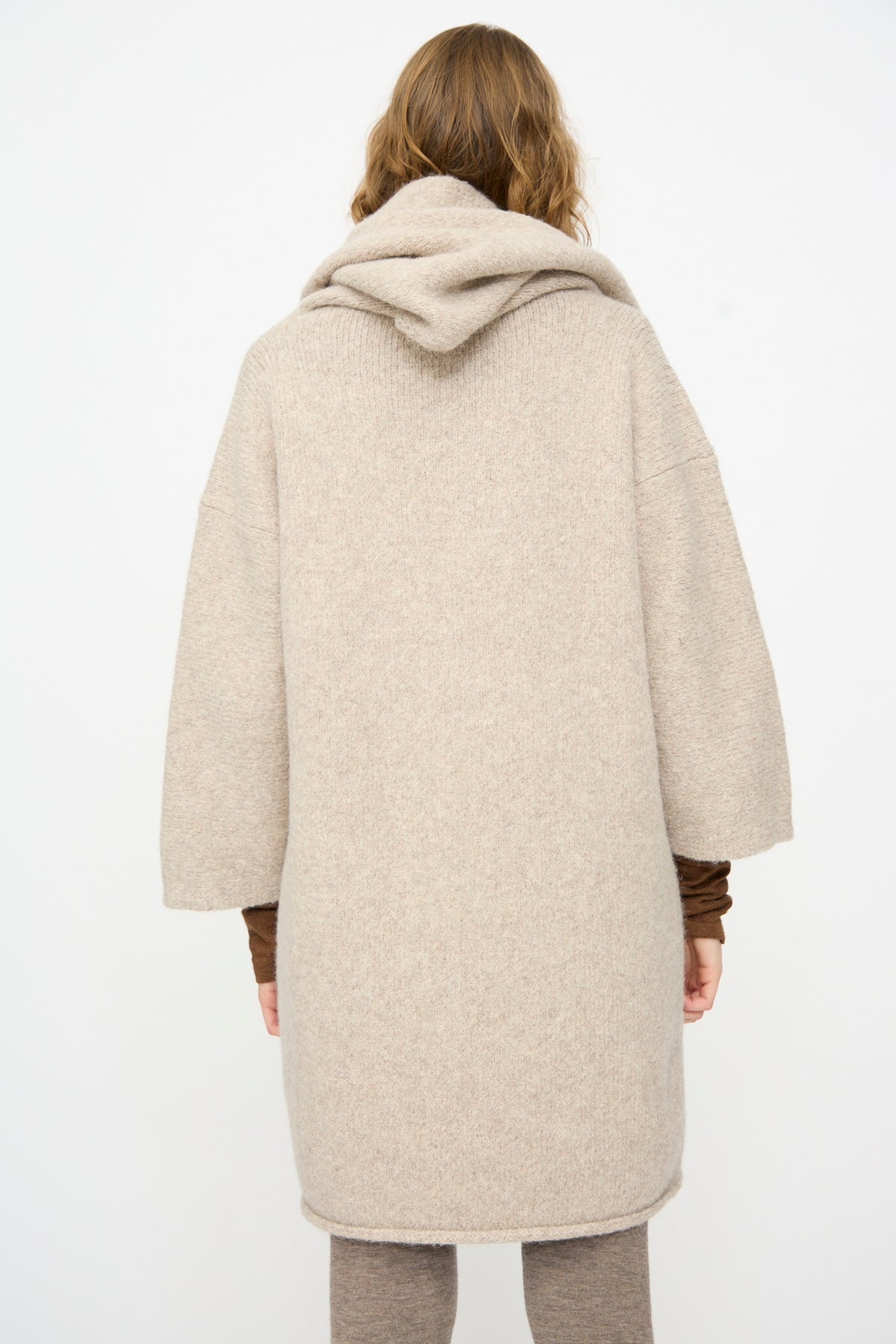 A person stands facing away, wearing the Lauren Manoogian Capote Coat in Pebble—a long, hand-loomed alpaca wool coat with loose sleeves—over brown clothing, set against a plain white background.