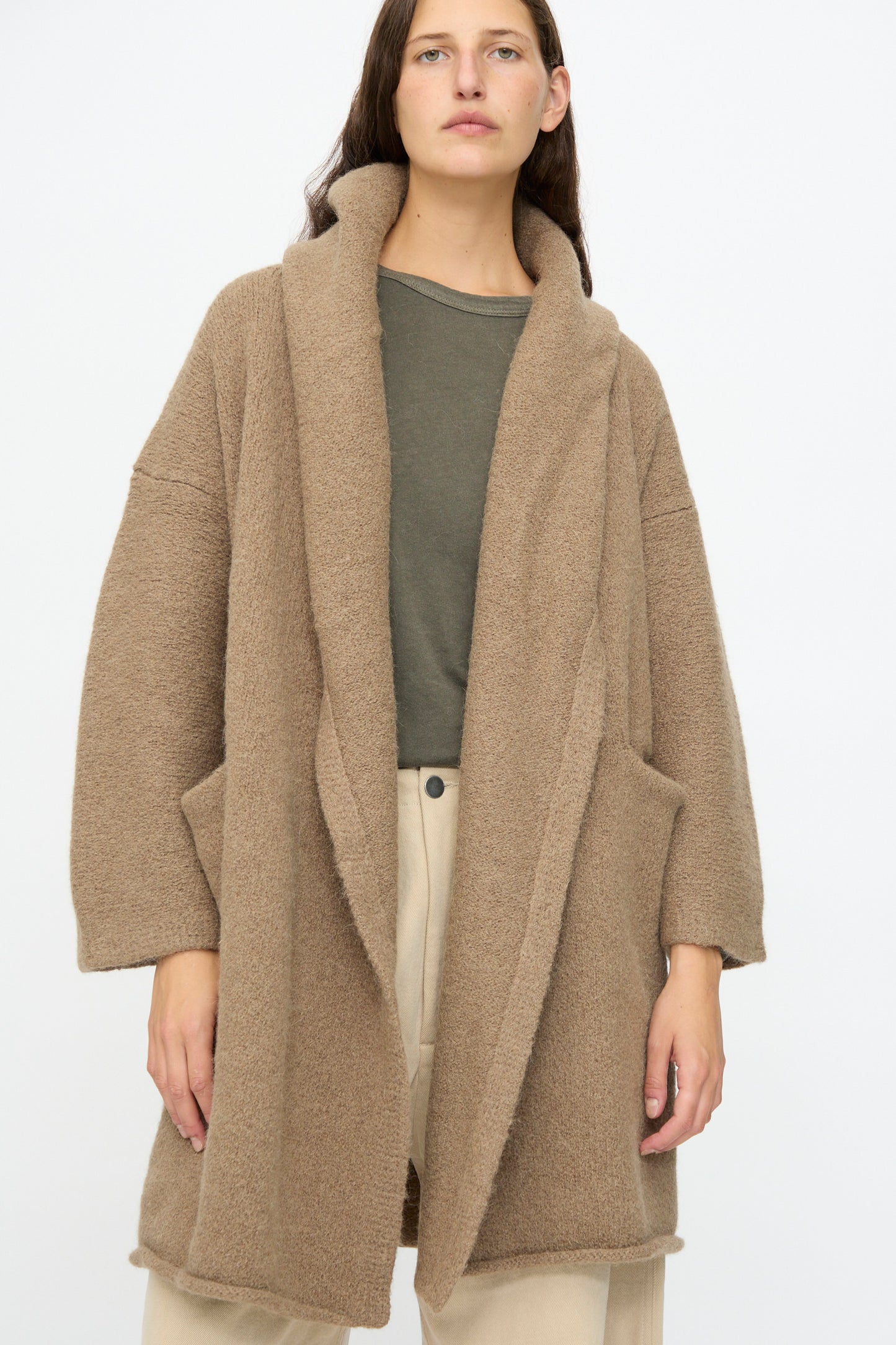 A person wears the Lauren Manoogian Capote Coat in Tobacco—a relaxed fit alpaca wool piece—over a green shirt and beige pants, standing against a plain light background.