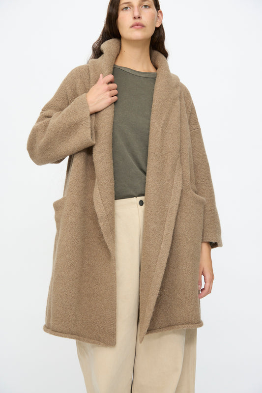 A person wears the Lauren Manoogian Capote Coat in Tobacco over a green shirt and beige pants, standing against a plain white background.