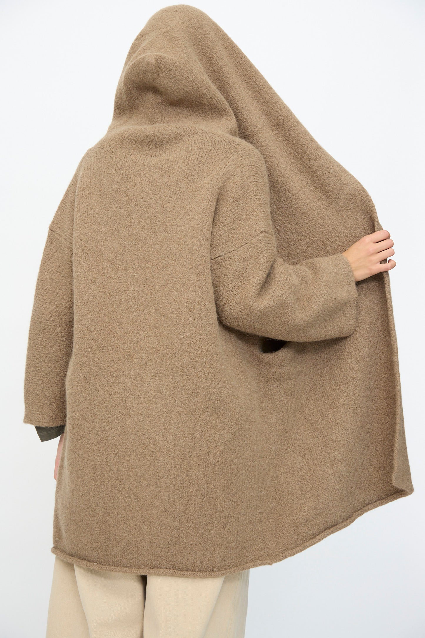 Model wearing the Capote Coat in Tobacco by Lauren Manoogian—a relaxed fit, tan hooded coat with the hood up—holds one side of the hood away from their face; back view shown against a plain background.