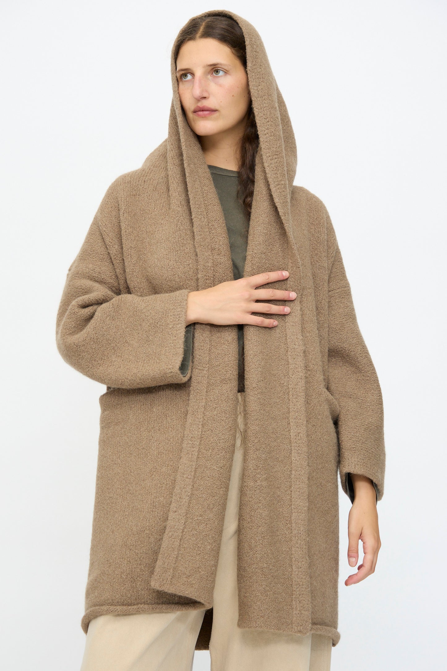 A woman wears the Lauren Manoogian Capote Coat in Tobacco—a relaxed fit brown hooded alpaca wool coat—over a green top and beige pants, standing against a plain white background.