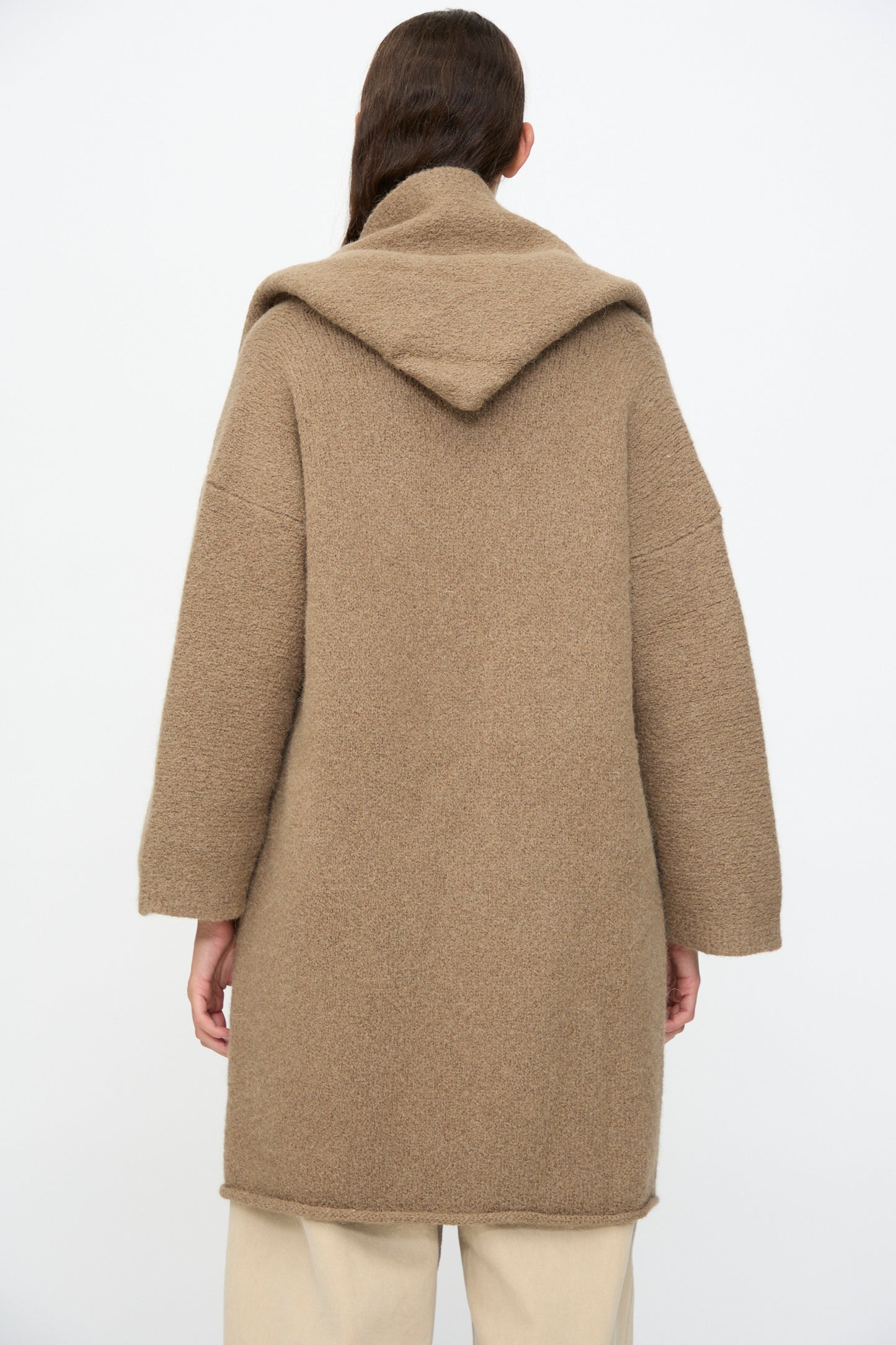 Person seen from the back wearing Lauren Manoogian's Capote Coat in Tobacco, a long, relaxed-fit hooded coat with loose sleeves, paired with light-colored pants against a plain white background.