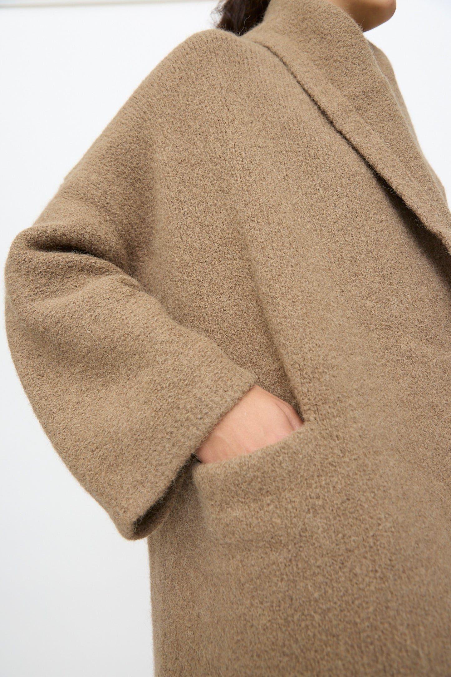 A person wearing the Lauren Manoogian Capote Coat in Tobacco, a textured light brown alpaca wool piece with a relaxed fit and one hand in the pocket, photographed from the side against a plain background.