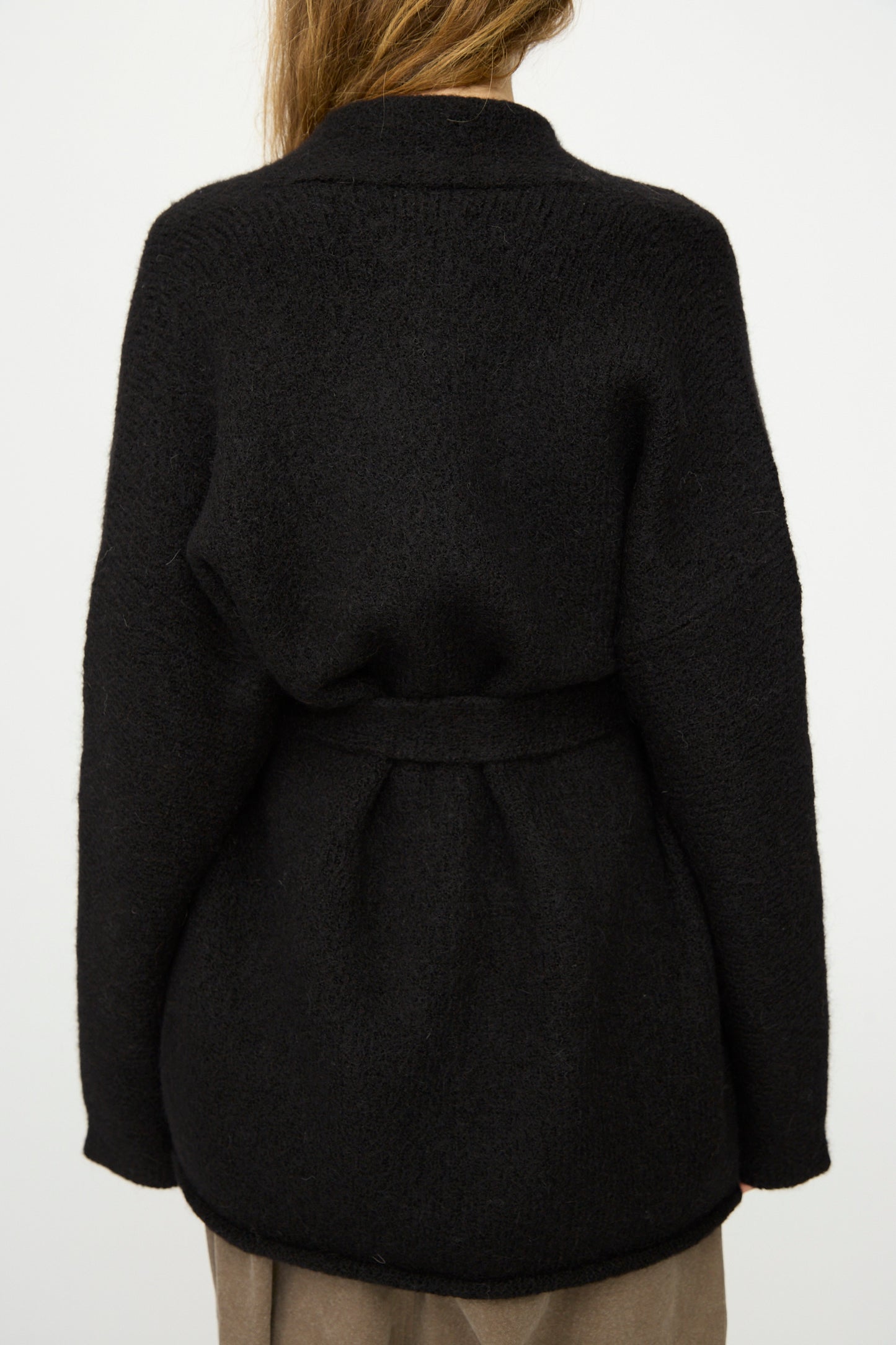 A person wearing the Lauren Manoogian Coto Cardigan in Black, seen from the back with long hair over one shoulder and brown pants.