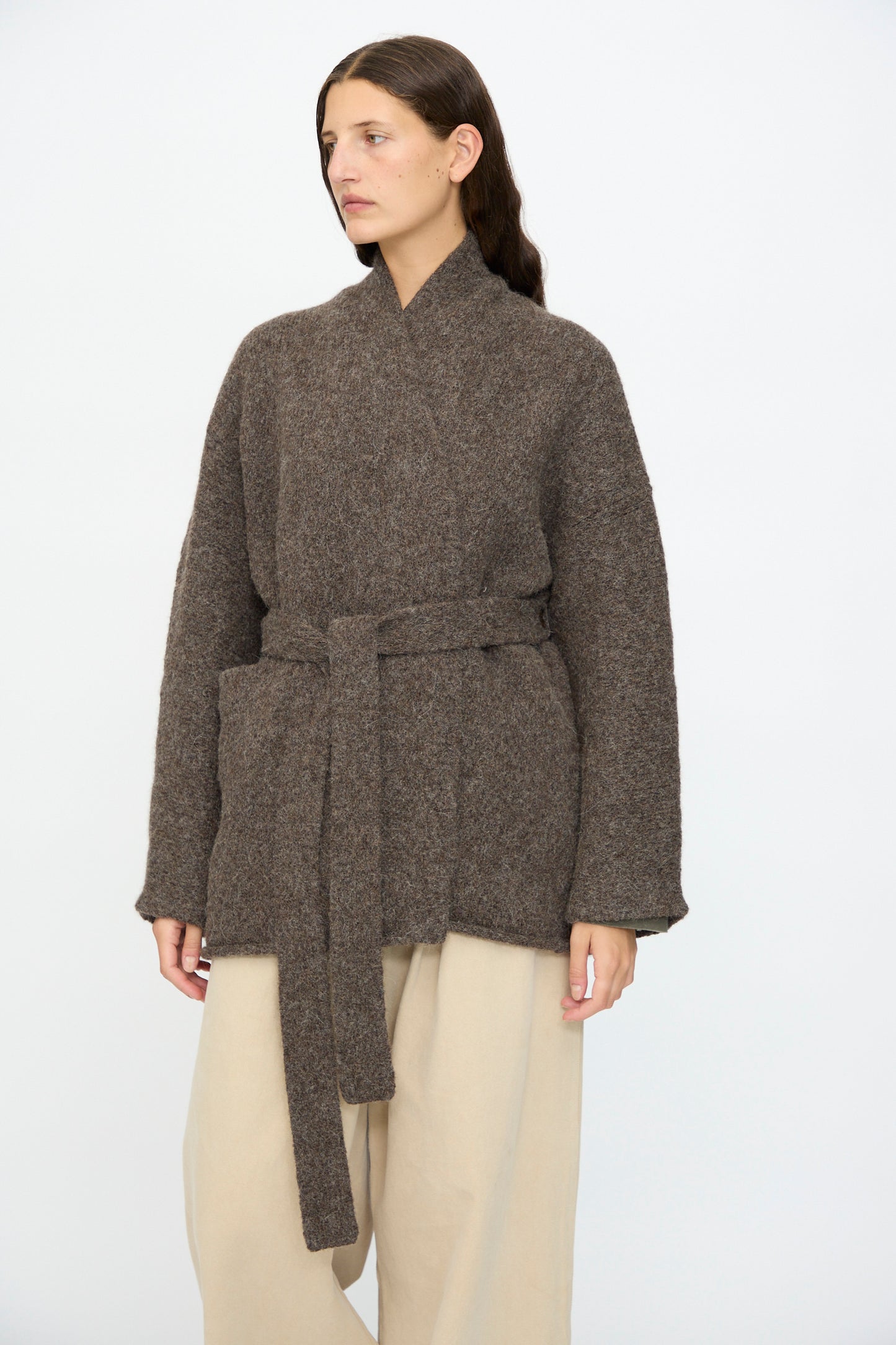 A woman stands against a plain white background wearing Lauren Manoogian’s Coto Cardigan in Patina, a hand-loomed alpaca piece with a relaxed fit, paired with beige wide-leg pants.