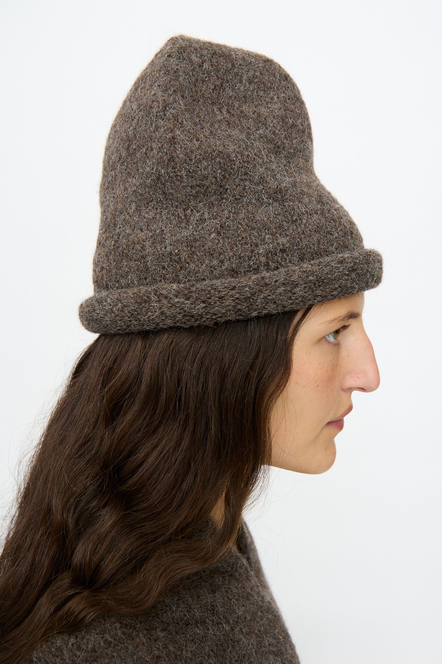 A person in profile wears the Lauren Manoogian Crown Beanie in Patina—a thick, textured, hand-loomed brown beanie with a broad folded brim—paired with a matching brown top, set against a plain white background.