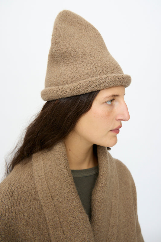 A person with long dark hair wears the Lauren Manoogian Crown Beanie in Tobacco, paired with a matching brown cardigan, shown in profile against a plain light background.
