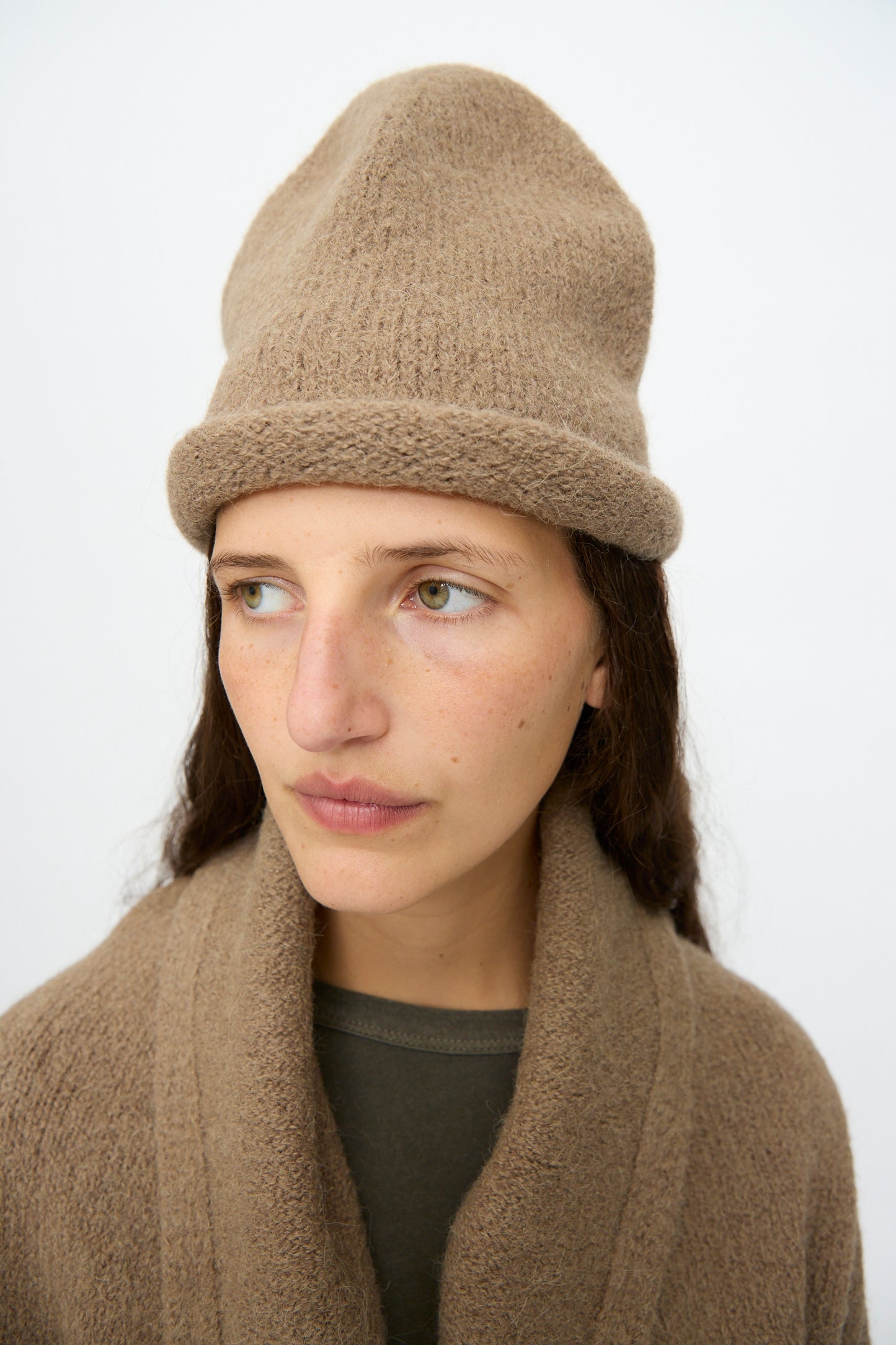 A person with long brown hair wears the Lauren Manoogian Crown Beanie in Tobacco, hand-loomed in Peru, and a matching cardigan over a dark shirt, looking slightly to the side against a plain background.
