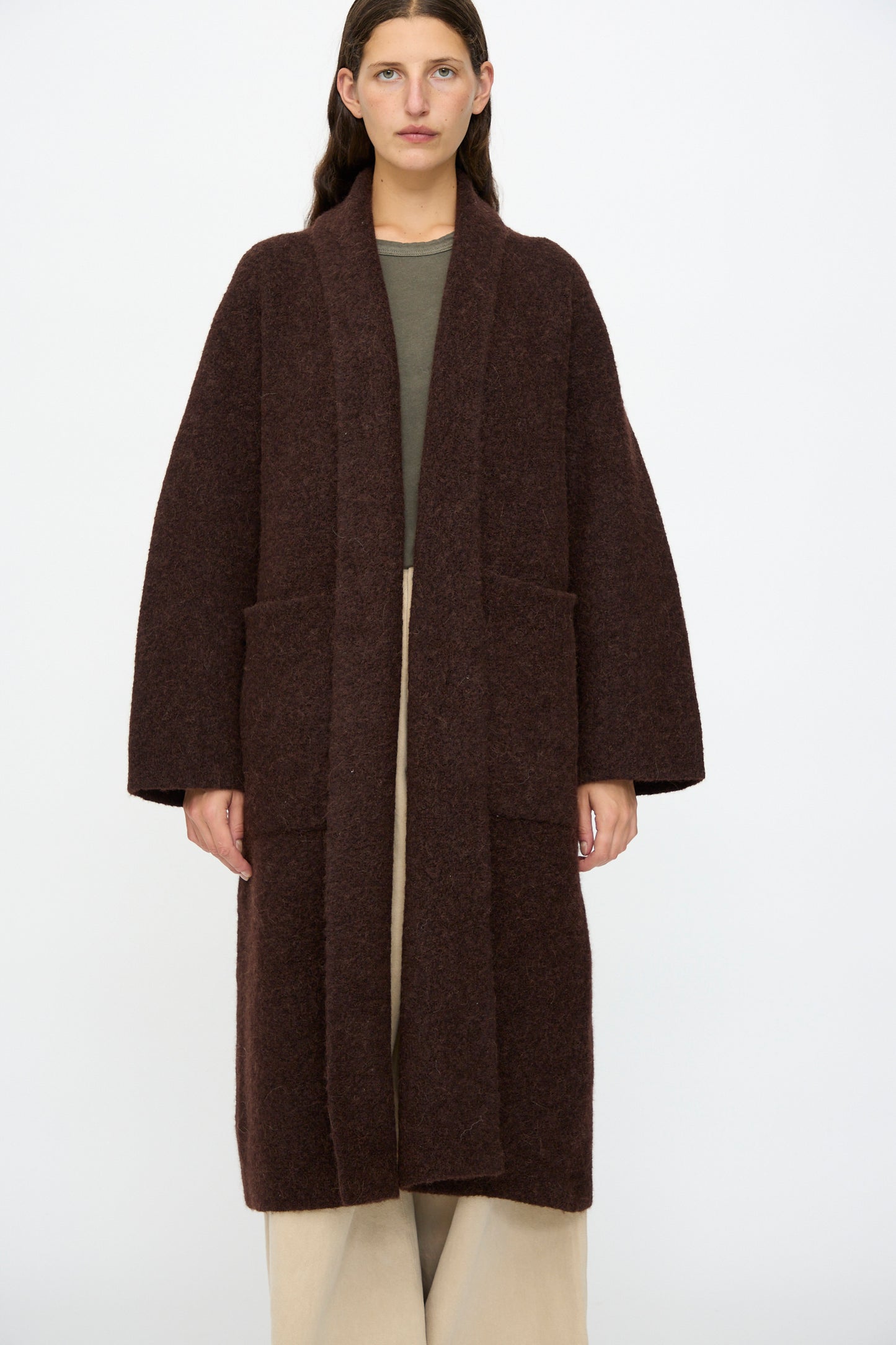 A person stands facing forward wearing the Lauren Manoogian Double Face Long Coat in Merlot over a green top and beige wide-leg pants, set against a plain white background.