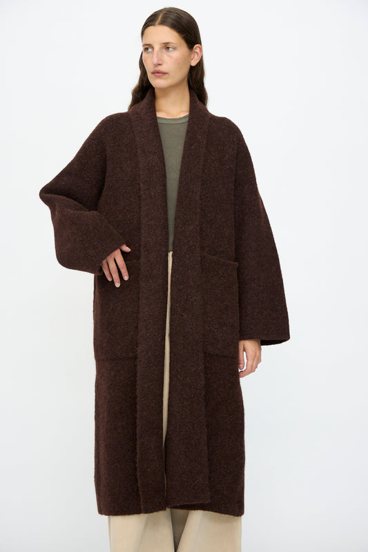 A person wears the Lauren Manoogian Double Face Long Coat in Merlot over a green top and beige pants, standing against a plain white background.