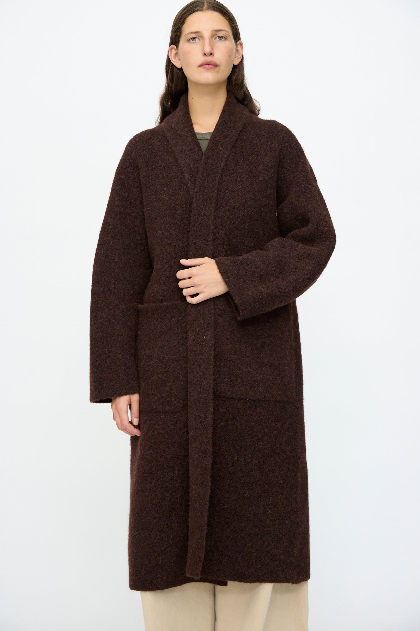 A person stands against a plain background wearing the Lauren Manoogian Double Face Long Coat in Merlot over beige pants, with one hand resting on the lapel.