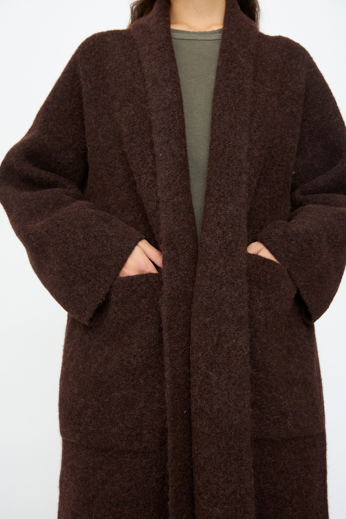 A person wearing Lauren Manoogian’s Double Face Long Coat in Merlot over a green top, with hands partially in the coat’s pockets.