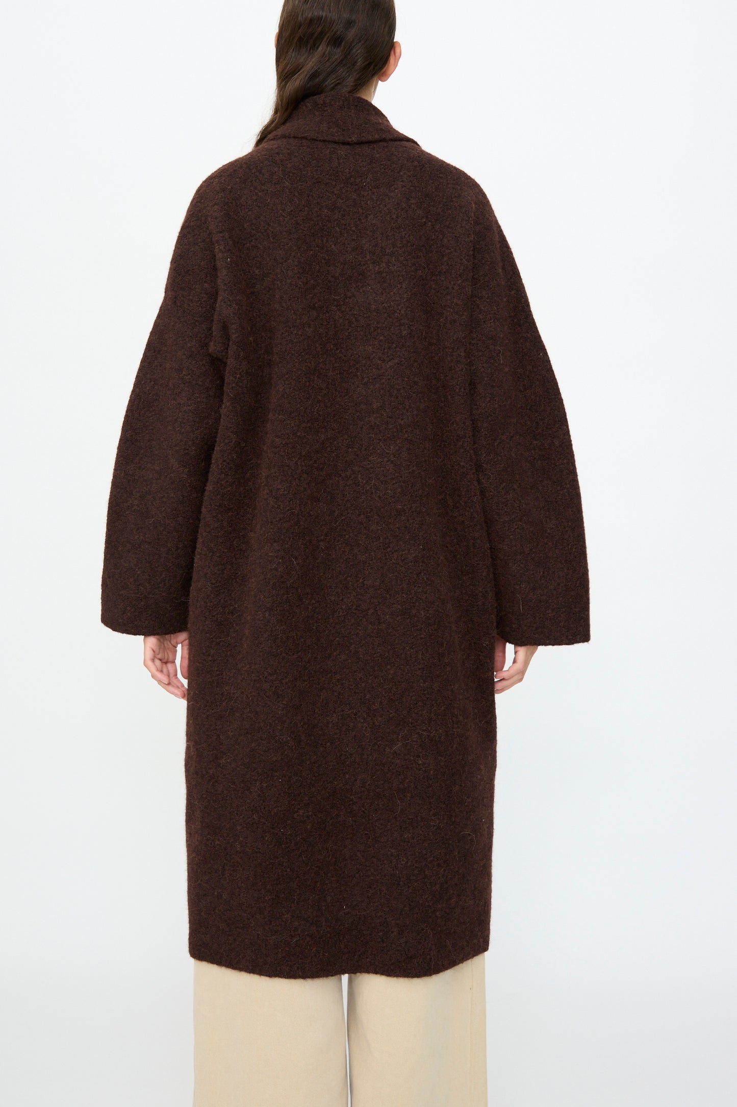 A person is shown from the back against a plain white background, wearing the Lauren Manoogian Double Face Long Coat in Merlot paired with light beige pants.