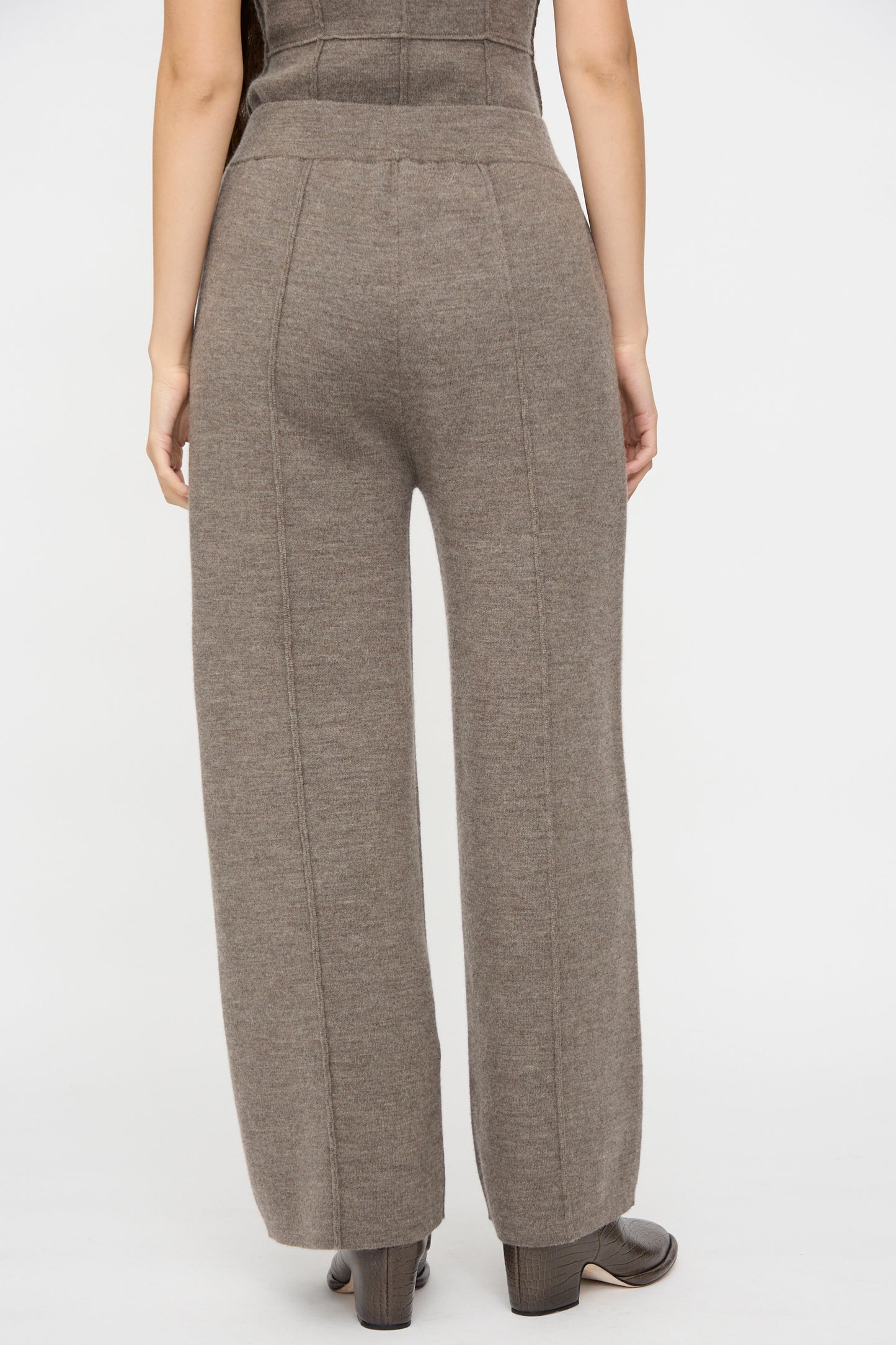 A person stands facing away in Lauren Manoogian’s Double Knit Trouser in Cinder—high-waisted, wide-leg taupe knit pants—paired with dark ankle boots, set against a plain white background.