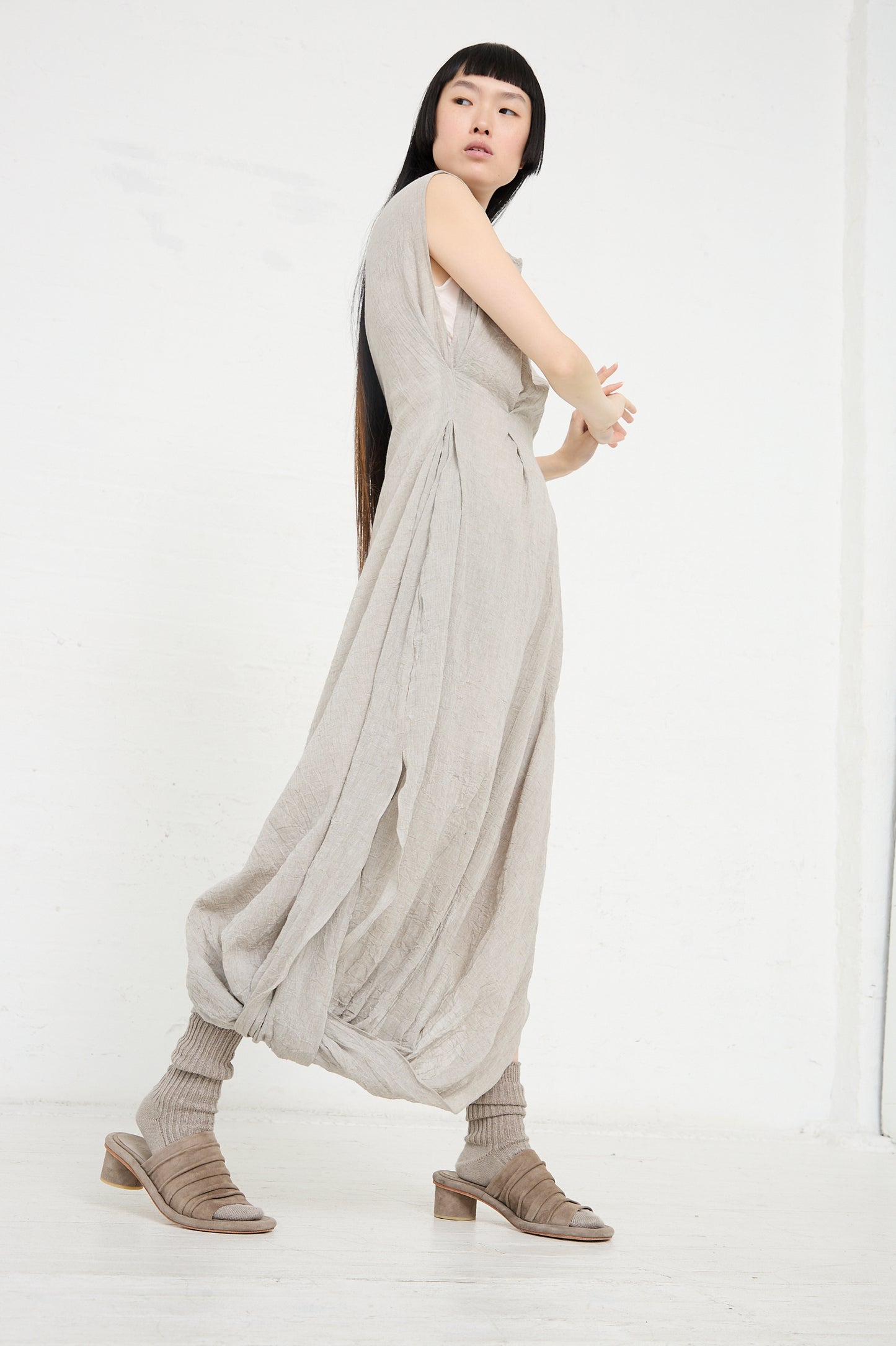 A woman in a flowing beige Lauren Manoogian Gauze Twist Dress in Ash and brown shoes poses in a white studio.