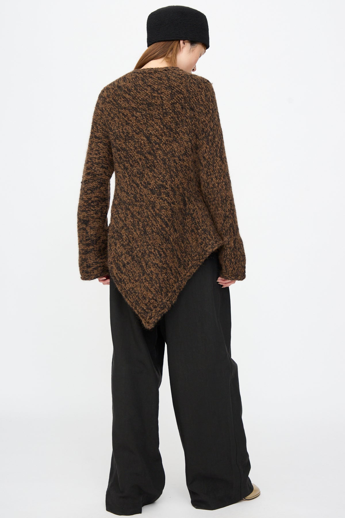 A person seen from the back wears a long, brown-and-black chunky alpaca Handknit Manta Pullover in Rust Marl with an asymmetrical hem and wide-leg black trousers.