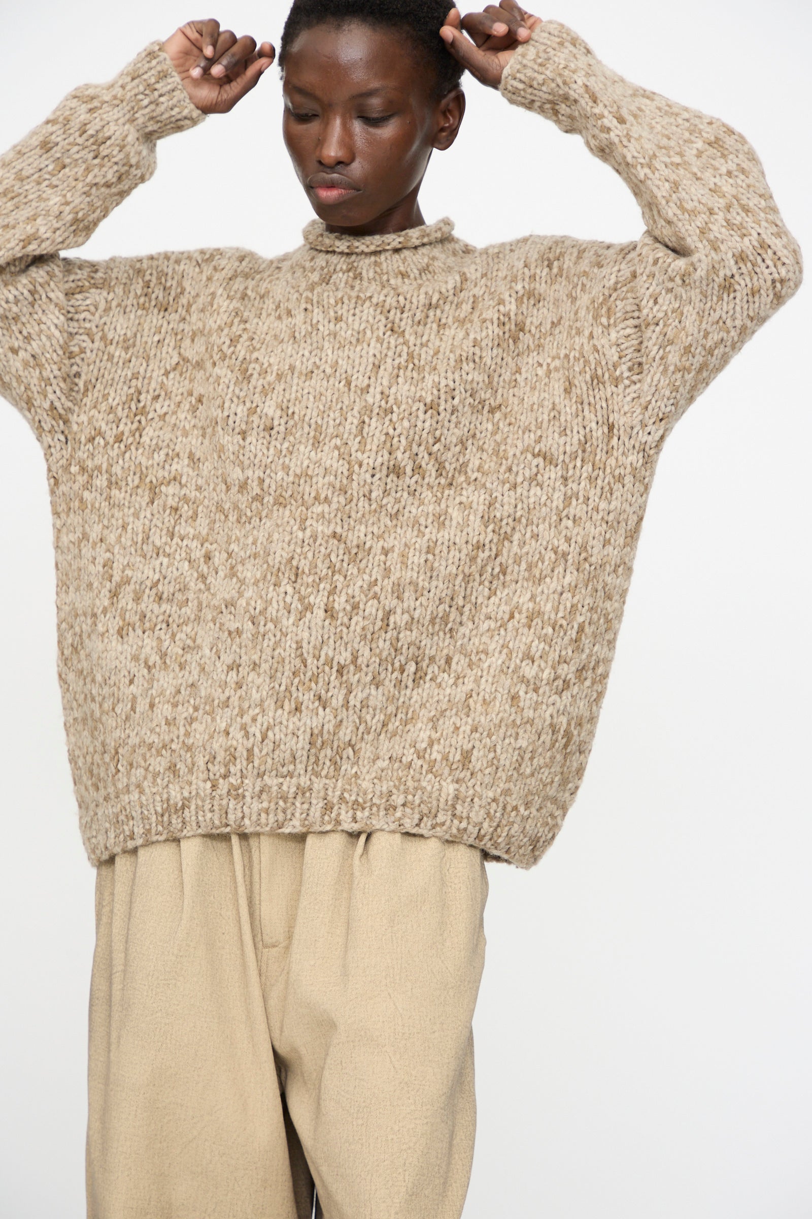 A person wearing the Lauren Manoogian Handknit Paint Rollneck in Tobacco and light beige pants stands with arms raised against a plain background.