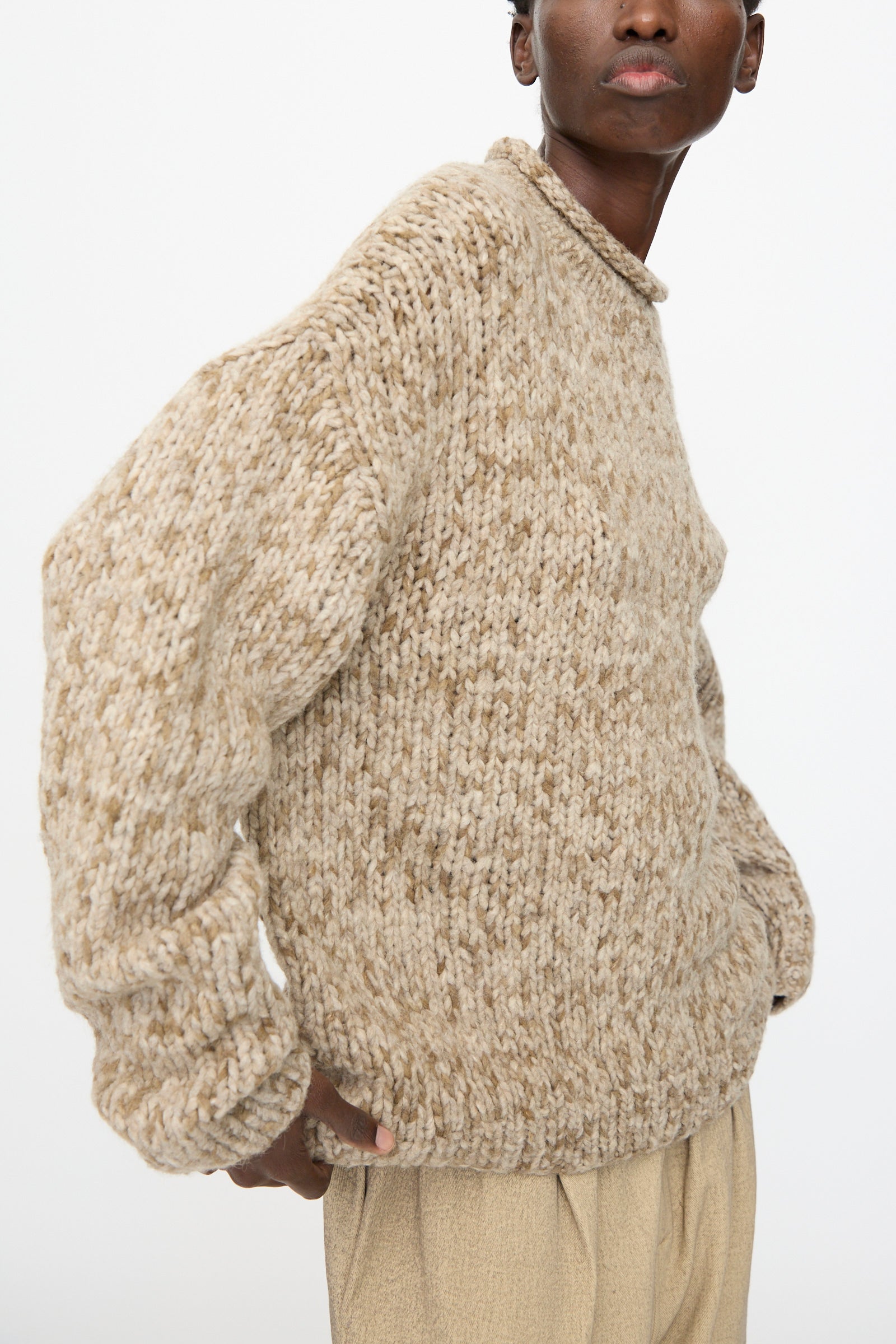 Person wearing the Lauren Manoogian Handknit Paint Rollneck in Tobacco, an oversized beige sweater handknit in Peru from soft baby alpaca, paired with light tan pants against a plain white background.
