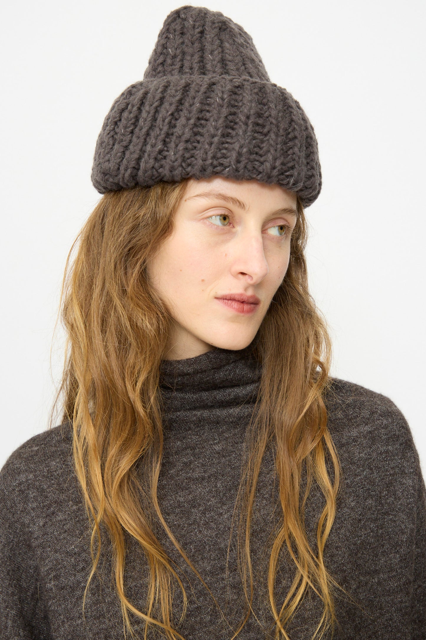A person with long, wavy hair wears the Lauren Manoogian Handknit Rib Hat in Coal and a dark turtleneck sweater, looking slightly to the side against a plain background.