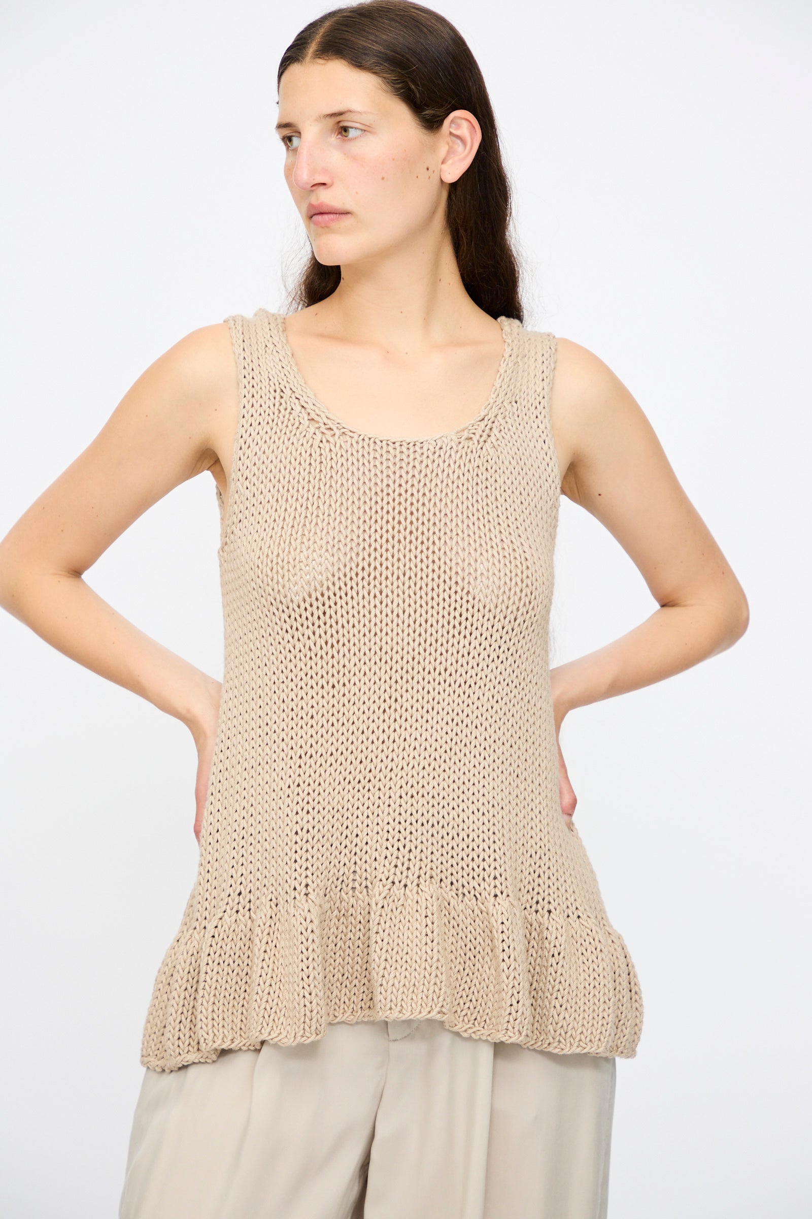 A woman wears the Lauren Manoogian Handknit Ruffle Tank in Sisal, a sleeveless beige pima cotton top with a ruffled hem, paired with light pants, standing against a plain white background.