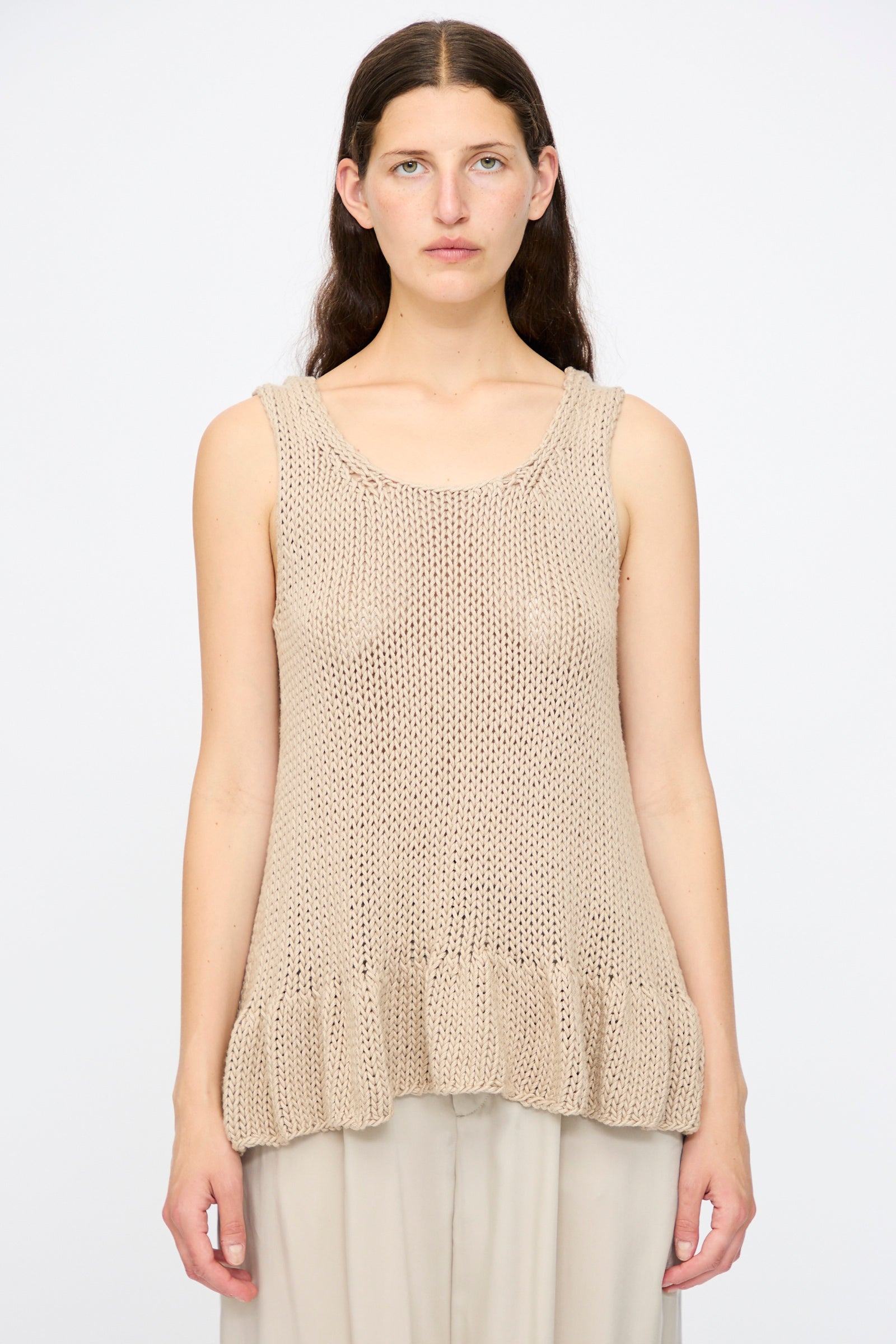 A person with long brown hair wears the Lauren Manoogian Handknit Ruffle Tank in Sisal, a relaxed-fit, sleeveless beige top with a ruffled hem, paired with light pants against a plain white background.