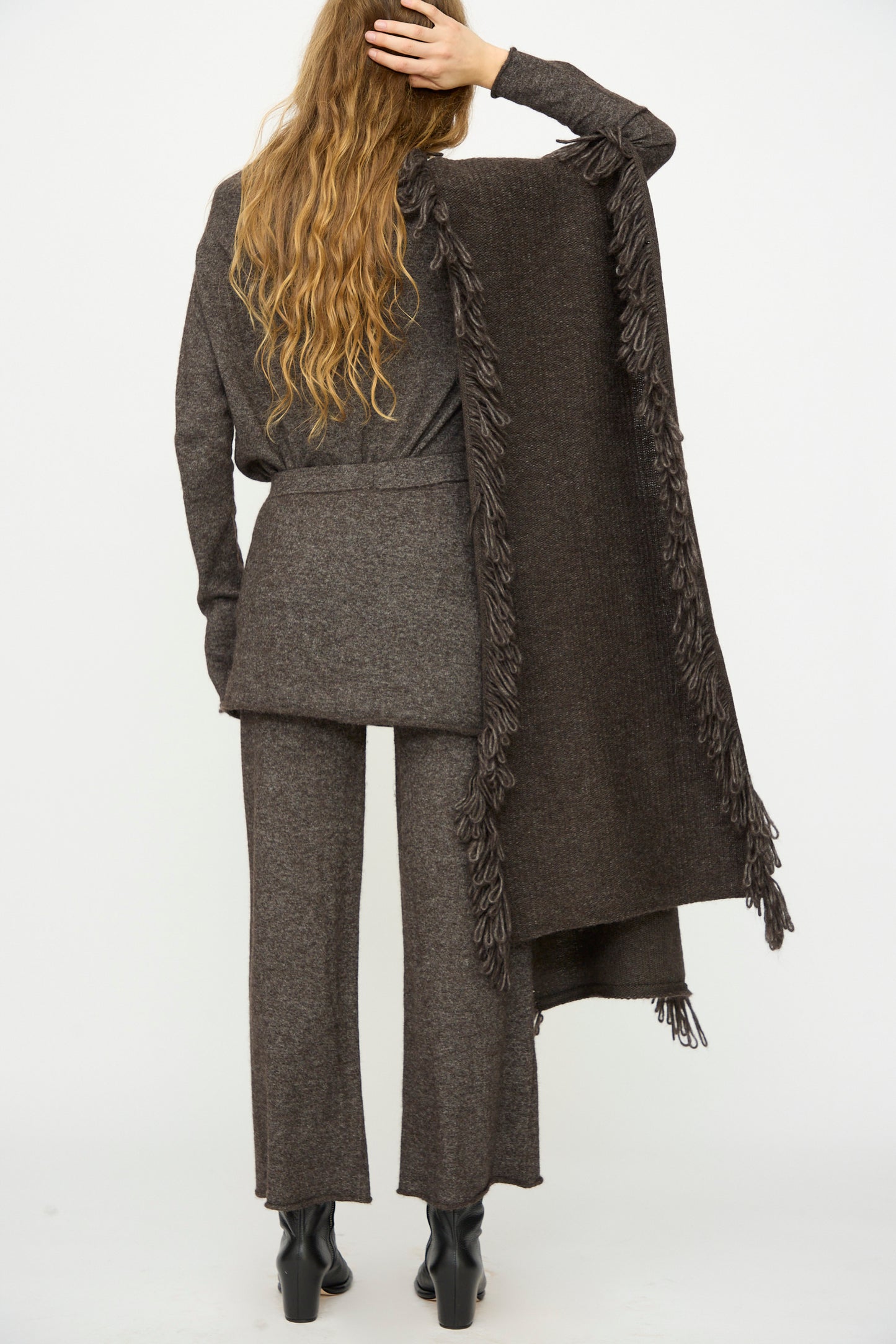 A person with long wavy hair stands facing away, wearing a brown knit outfit, black boots, and the Lauren Manoogian Knitweave Wrap in Blackened draped over one shoulder.
