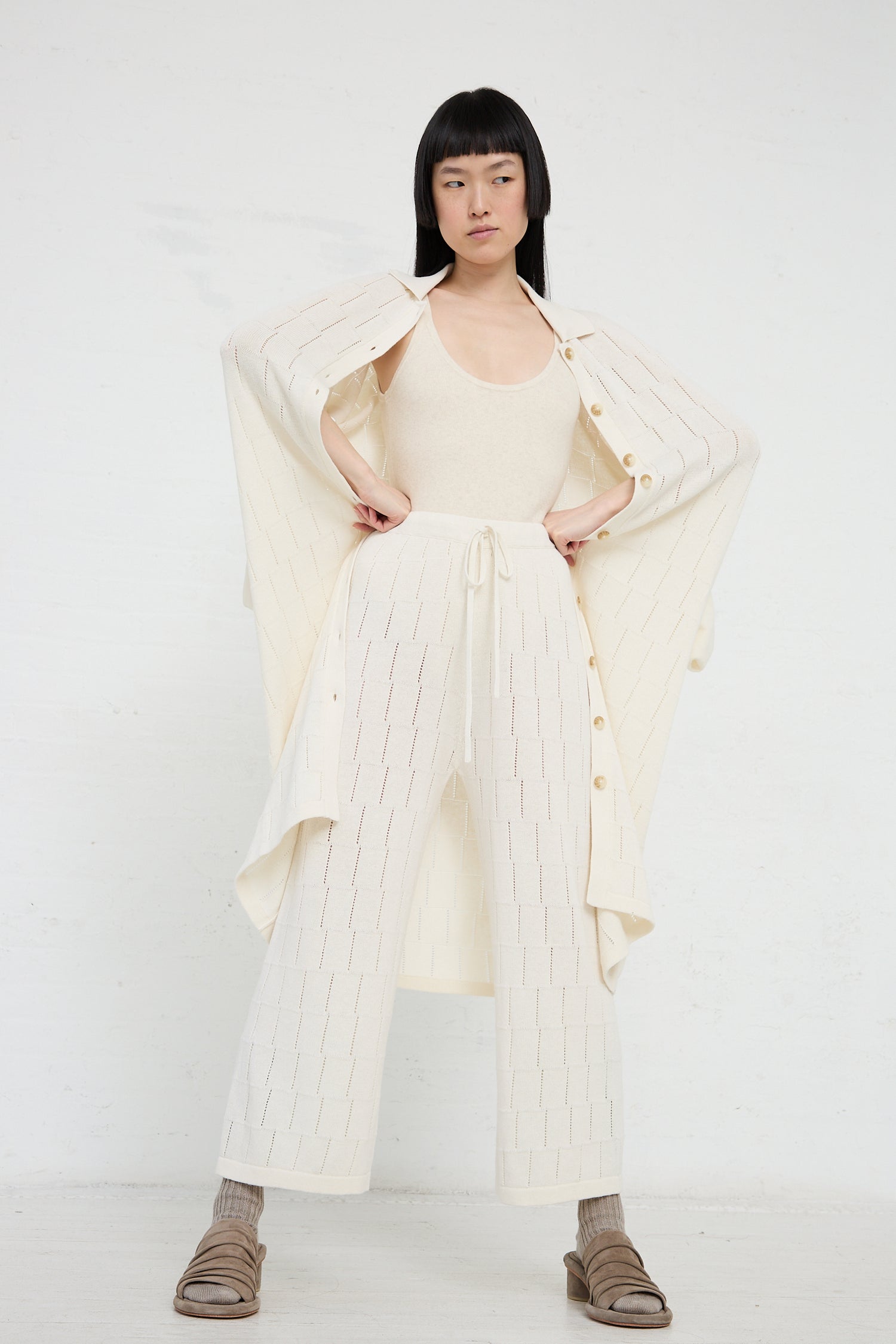 Woman in a cream-colored ensemble with trousers made from Lauren Manoogian's Lattice Pant in Bone, a sustainable pima cotton linen blend, and an open long jacket, posing with hands on hips against a white backdrop.
