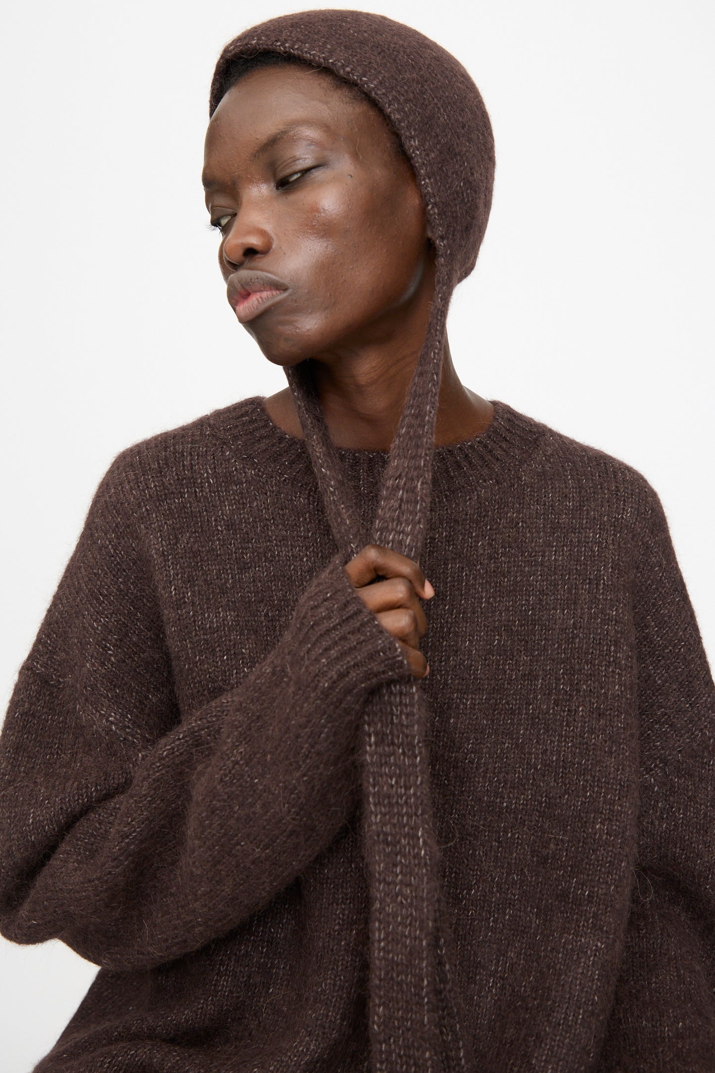 A person wearing the Lauren Manoogian Loft Bonnet in Soil, holding one strap with a neutral expression, against a plain light background. Made in Peru.
