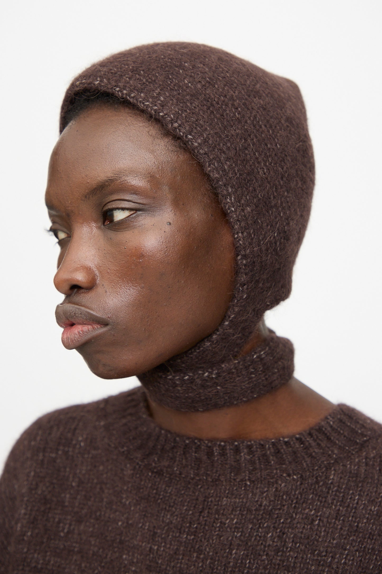 A person wears the Lauren Manoogian Loft Bonnet in Soil, a brown suri alpaca blend knit balaclava, paired with a matching sweater, shown in profile against a plain white background.