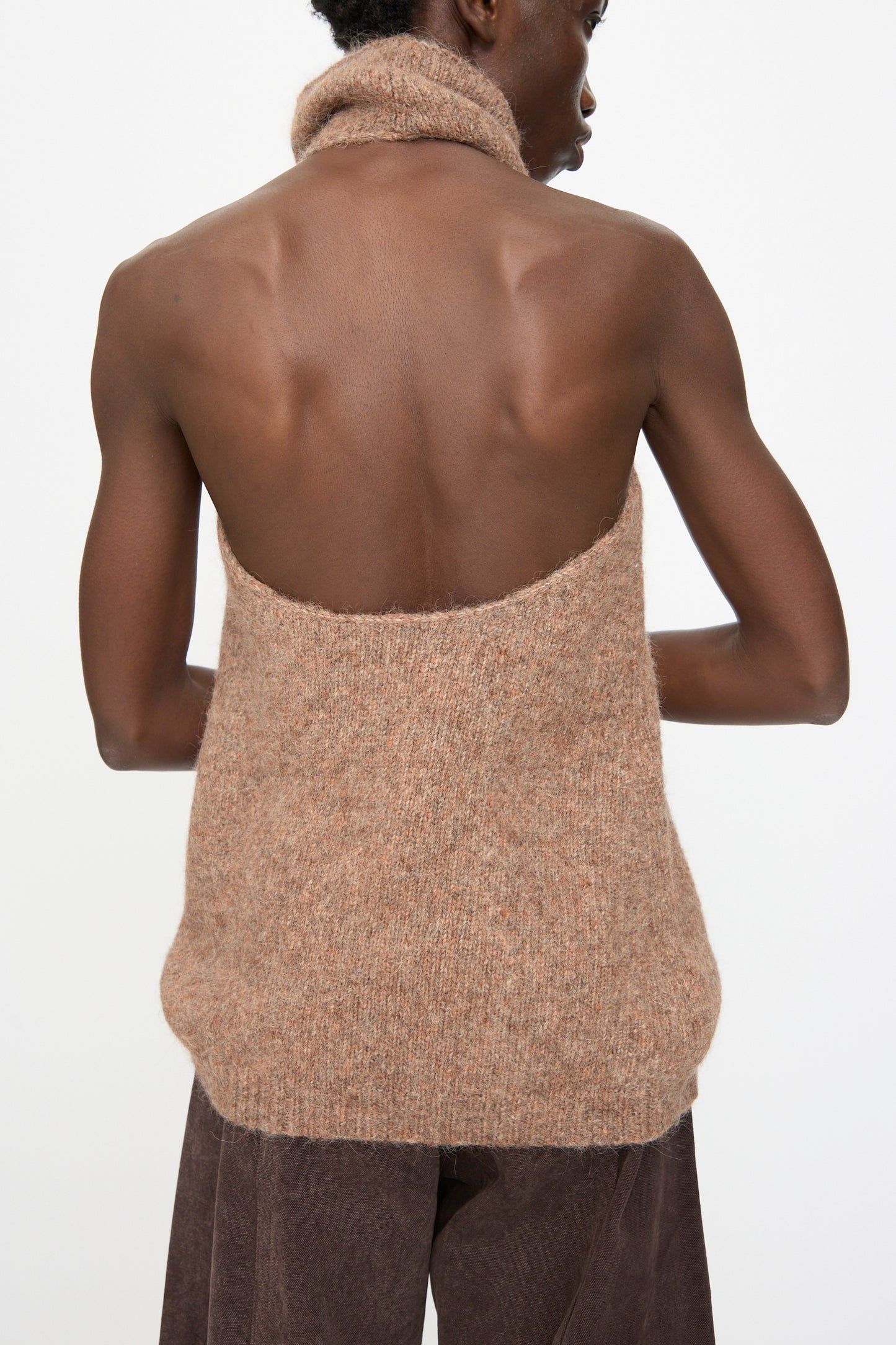 A person wears the Lauren Manoogian Loft Halter in Spice—an alpaca blend sleeveless turtleneck with a ribbed hem—paired with brown pants, viewed from the back against a plain white background.