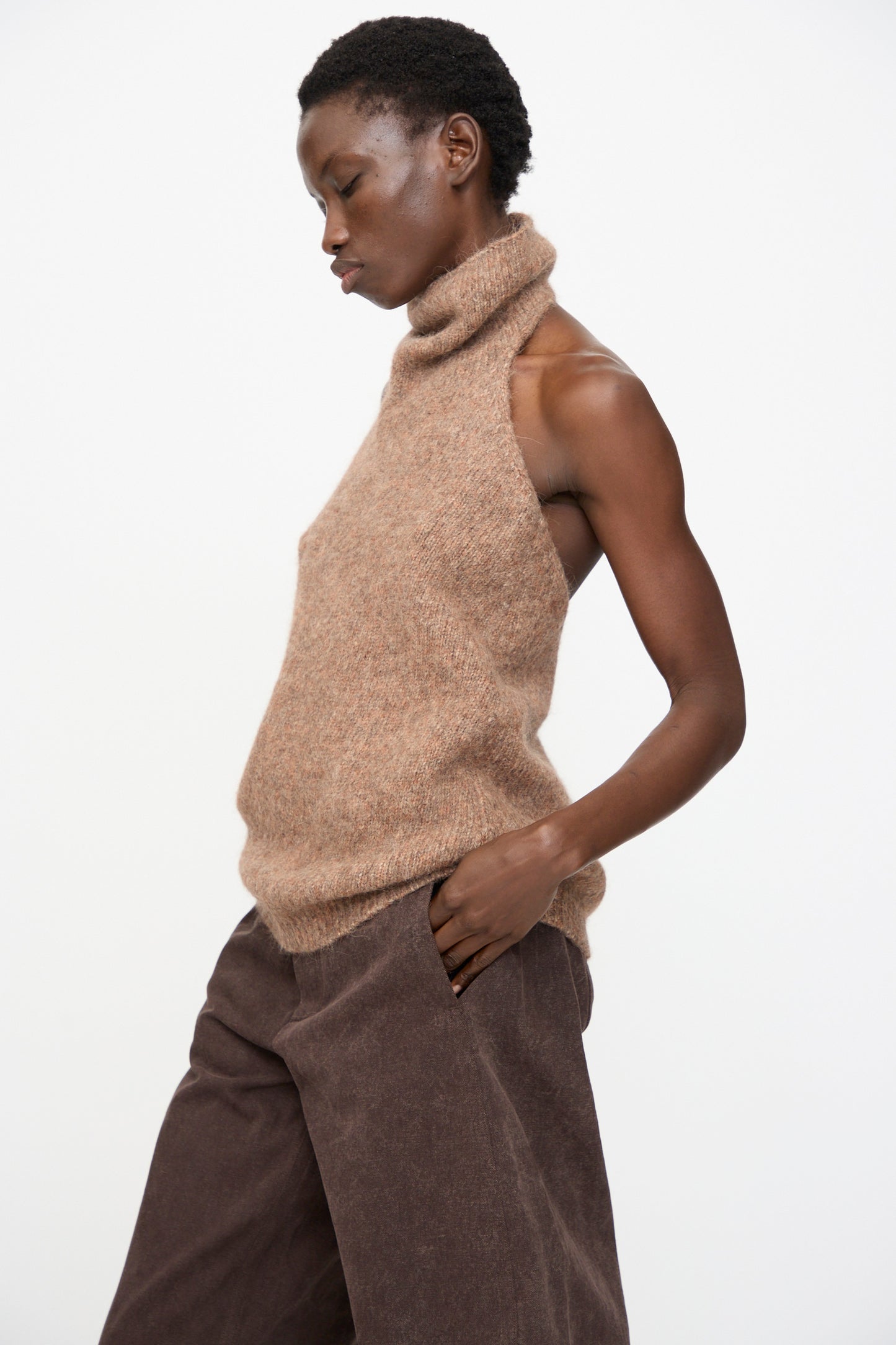 A person stands in profile against a plain white background, wearing the Lauren Manoogian Loft Halter in Spice—a sleeveless alpaca-blend turtleneck top with ribbed hem—paired with wide-leg brown pants.