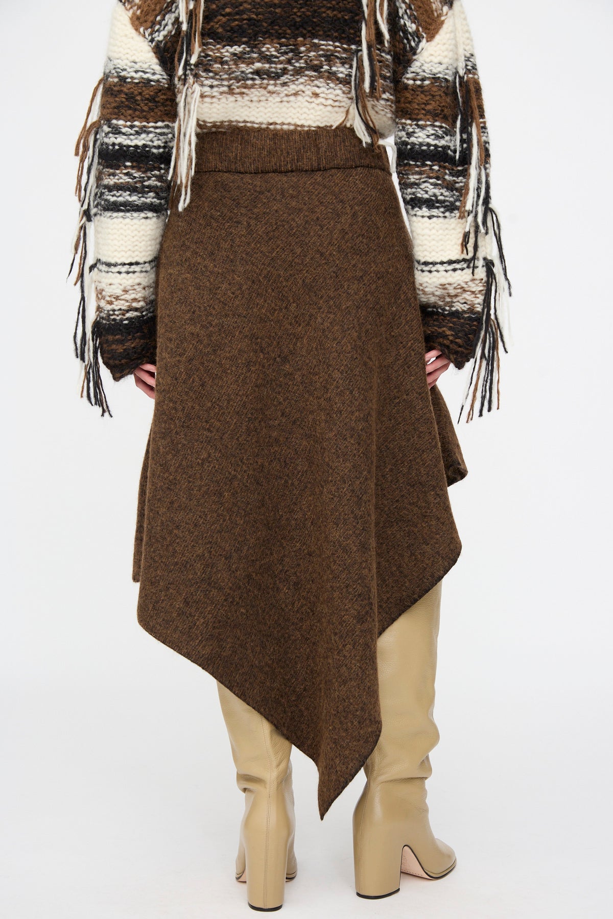 Back view of the Lauren Manoogian Manta Skirt in Rust Marl, a brown, mid-length merino wool skirt with an asymmetrical hem, worn with a fringed, multi-colored knit sweater and tan leather boots.