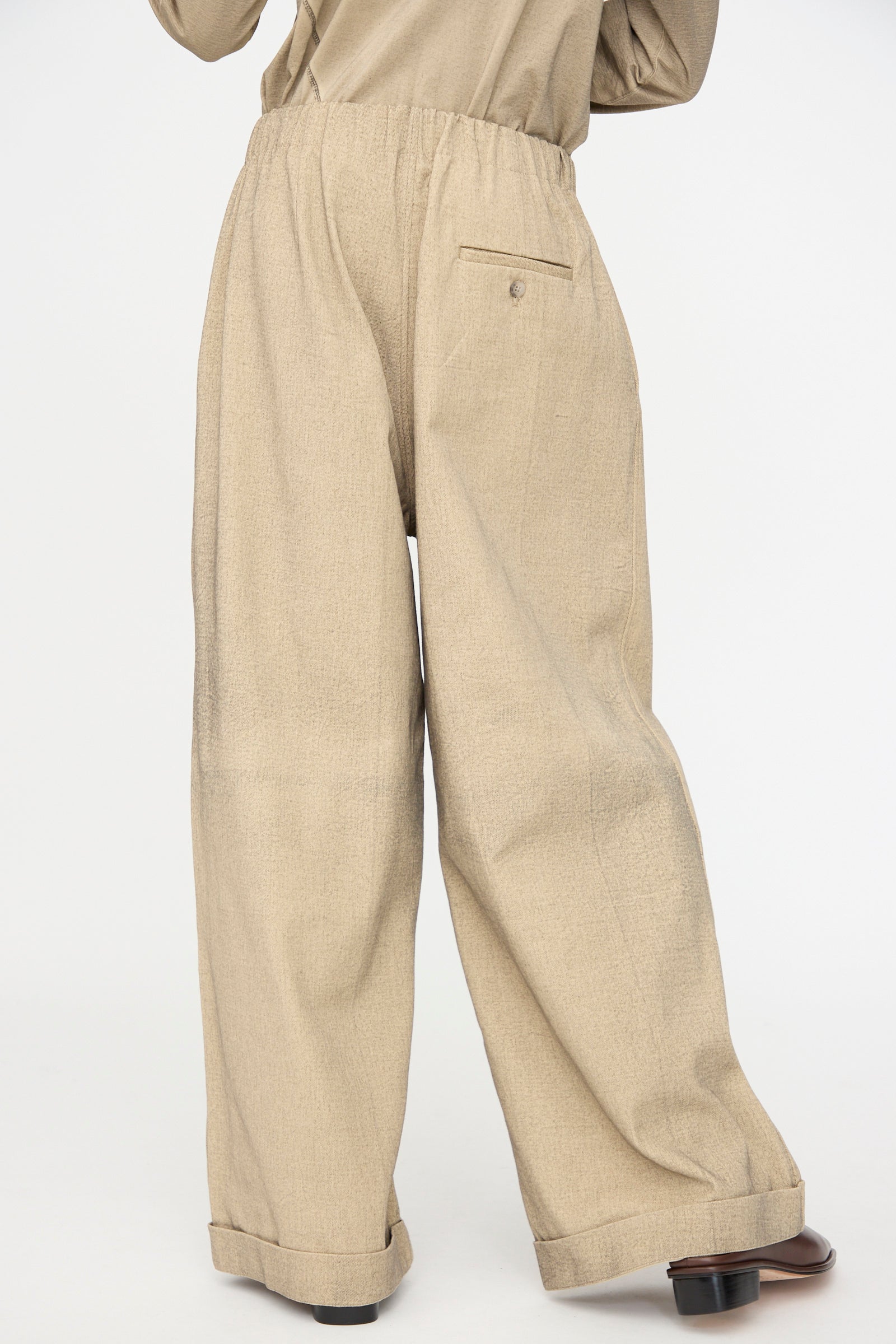 Person wearing Lauren Manoogian Painted Trouser in Antique—loose-fitting, high-waisted cotton pants with a back pocket and cuffed hems—paired with black shoes, shown from behind on a plain white background.