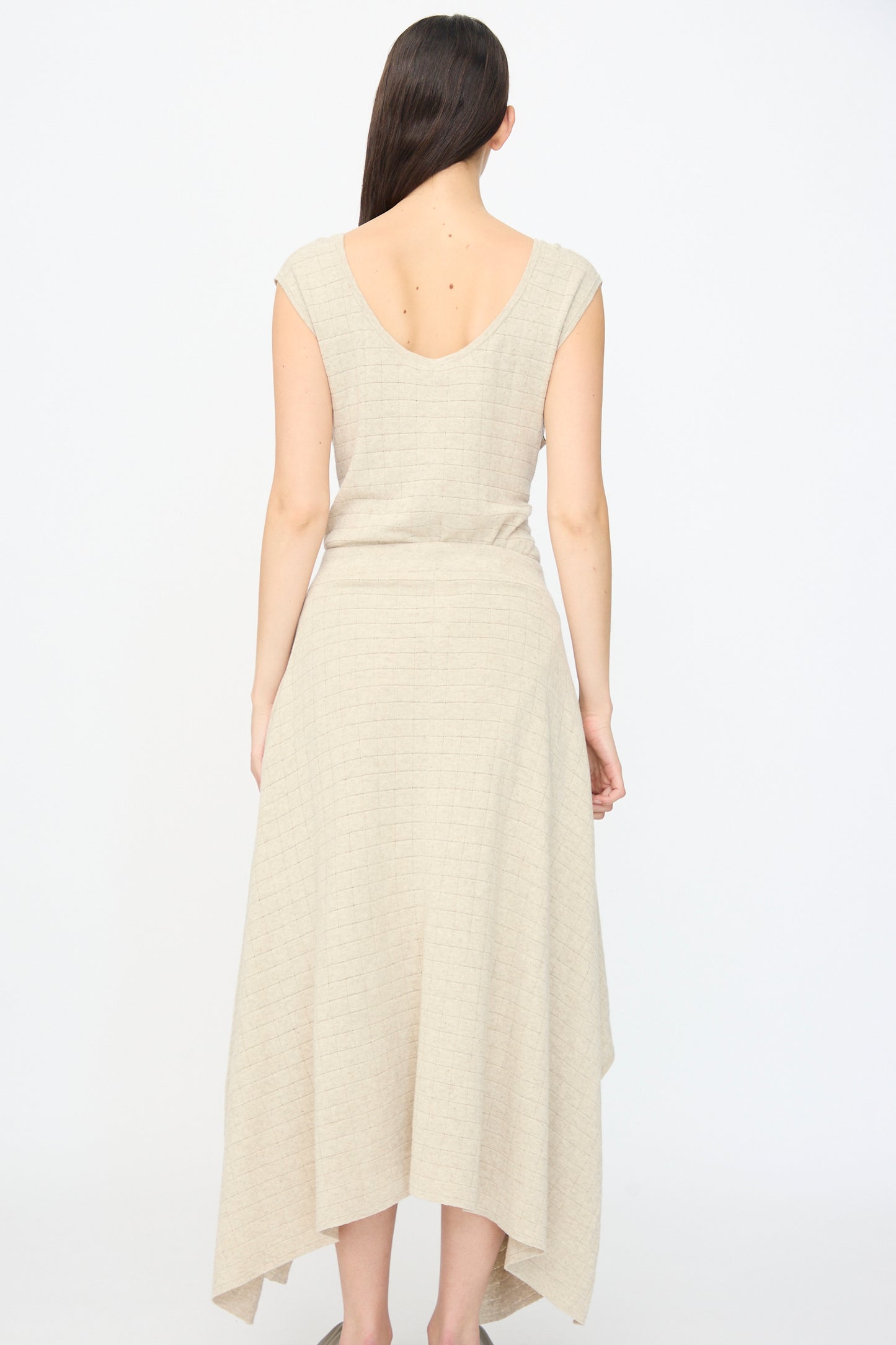 A woman stands with her back to the camera, wearing the Lauren Manoogian Planes Skirt in Flax, a sleeveless beige dress with a handkerchief hem, set against a plain white background.