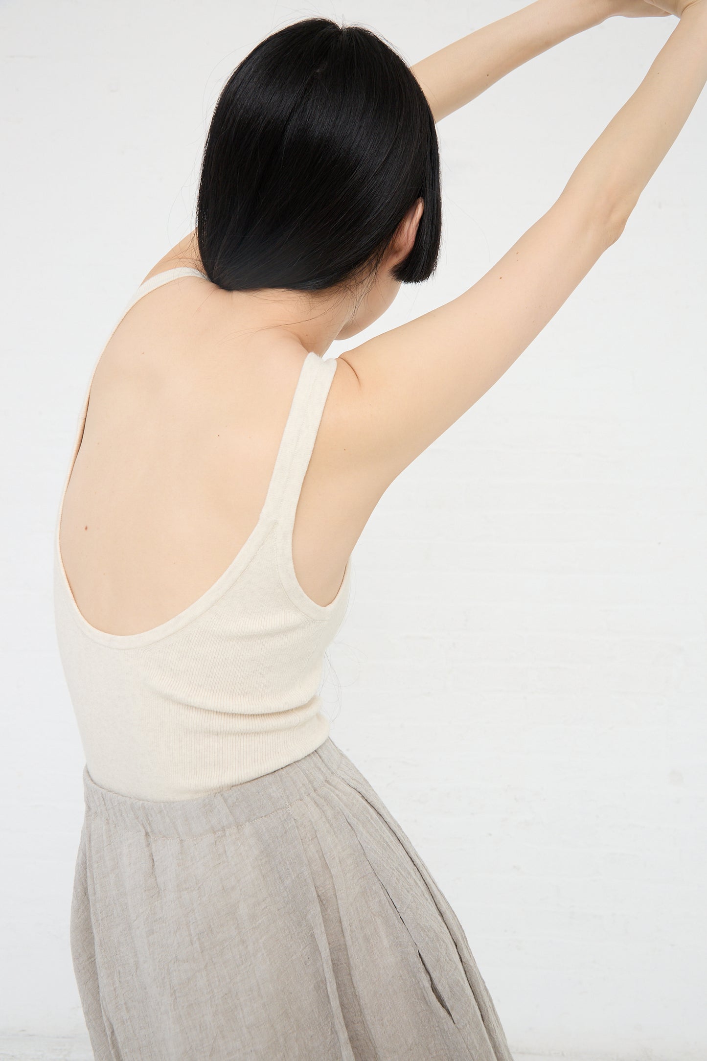 A woman with her back turned, reaching up with one arm, wearing a Rib Bodysuit in Hessian and skirt against a white background by Lauren Manoogian.