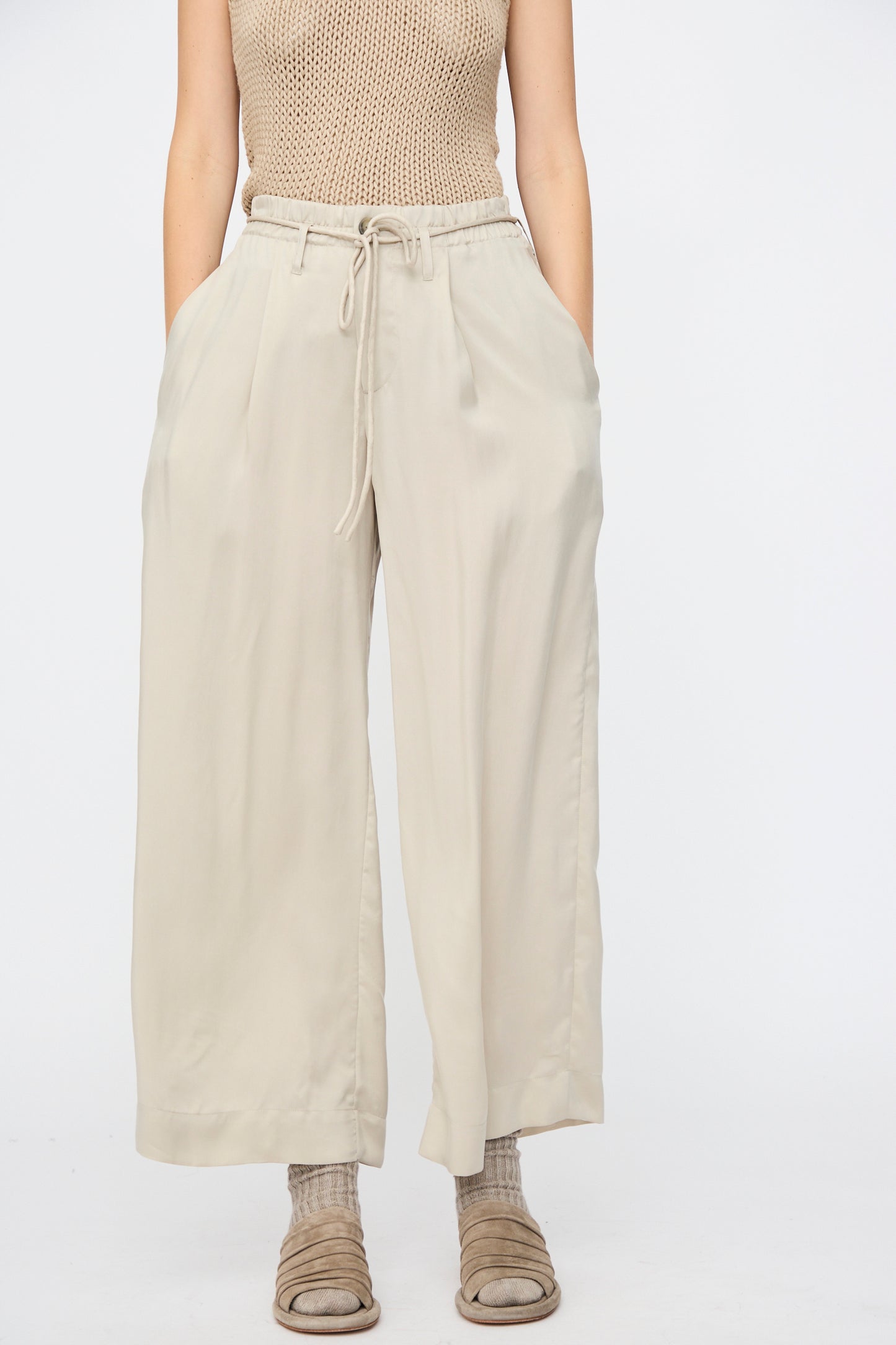 Wearing a sleeveless beige knit top, Lauren Manoogian’s Sanded Trouser in Sisal—high-waisted, wide-leg drawstring pants—and beige sandals with socks, this person stands against a plain background.