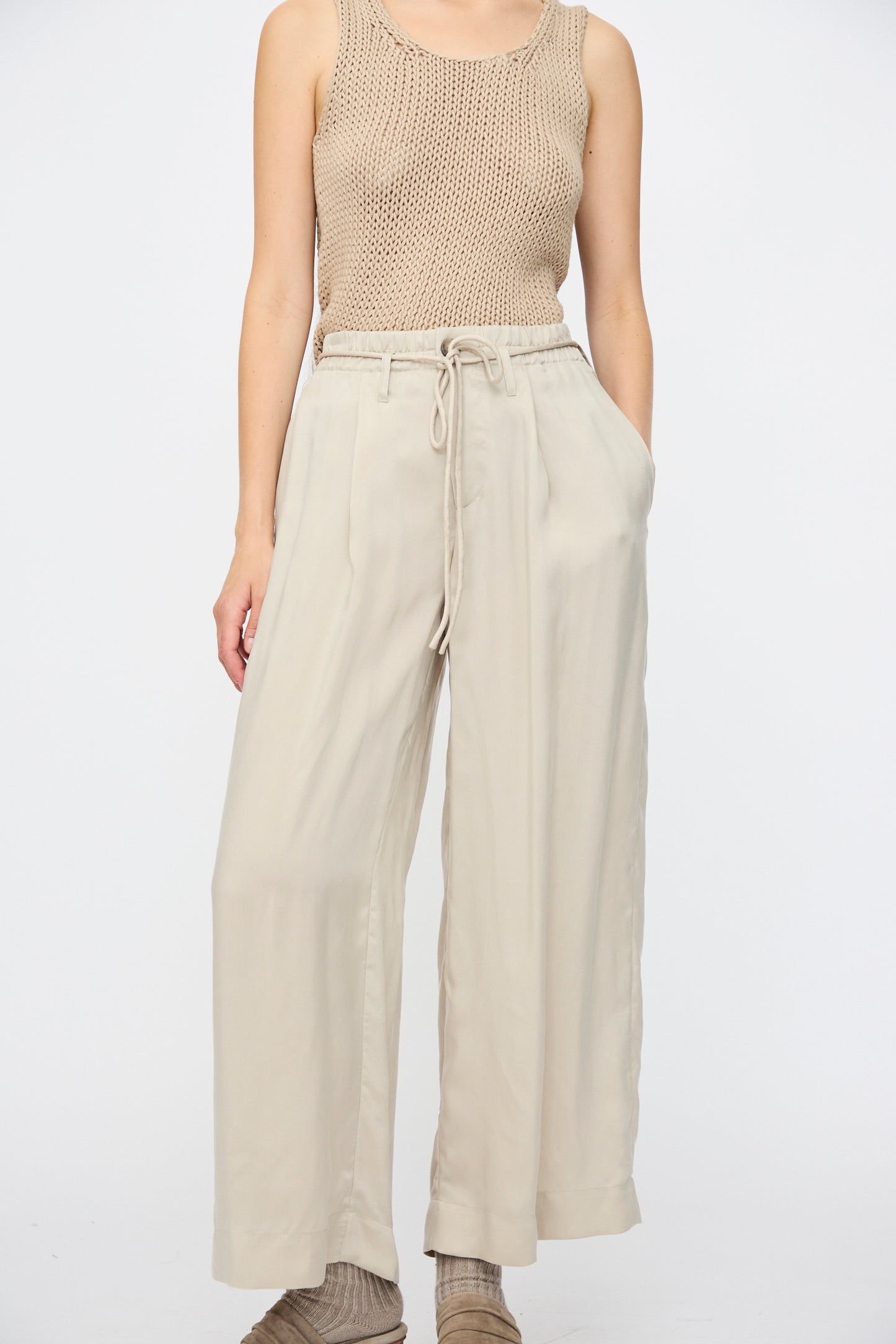 A person wears the Lauren Manoogian Sanded Trouser in Sisal—loose, high-waisted wide leg drawstring pants—with hands in pockets, and a sleeveless beige knit top, standing against a plain white background.