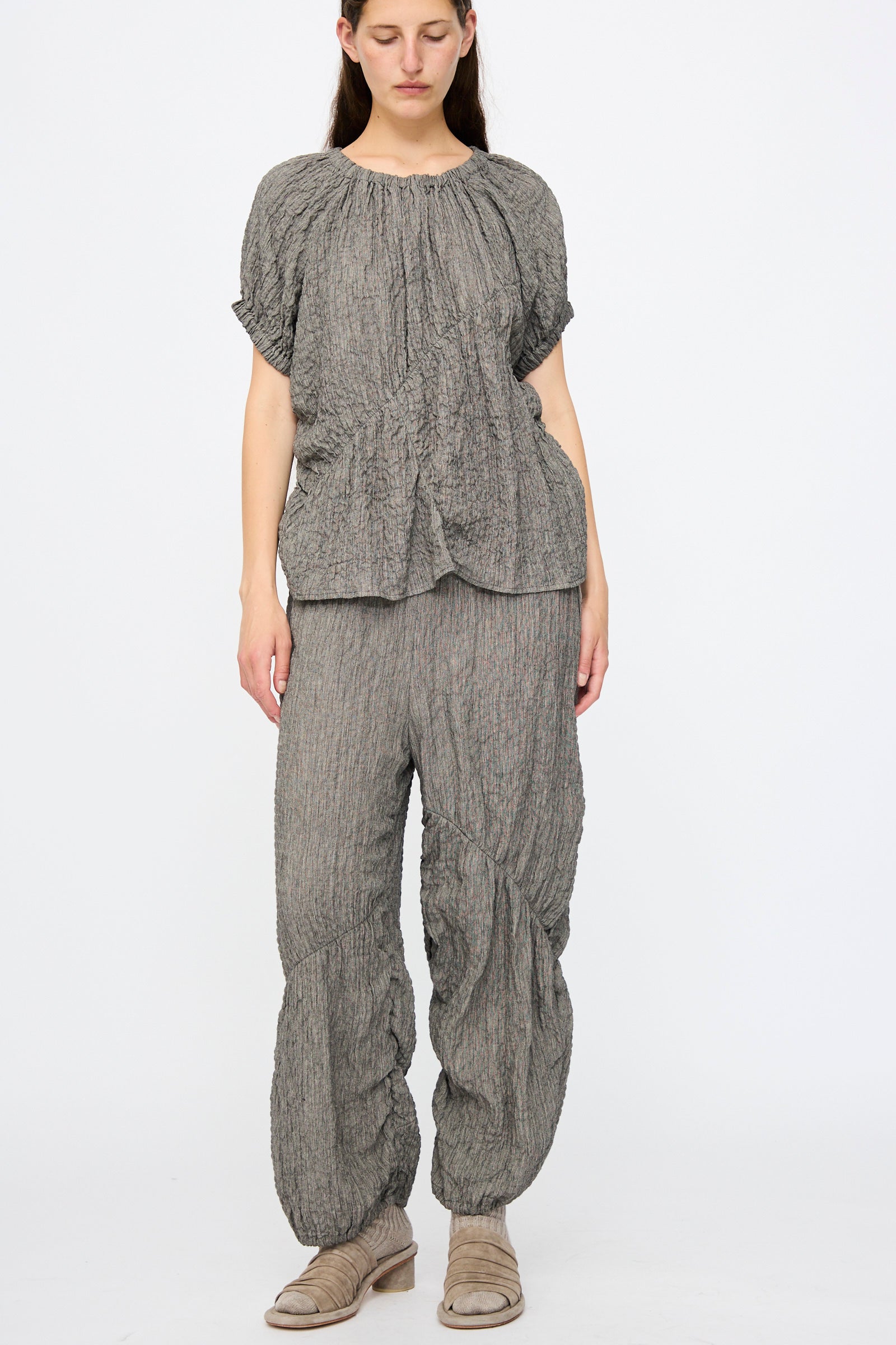 A woman stands against a plain background wearing Lauren Manoogian Shirred Pants in Charcoal, featuring a relaxed fit and elasticated waist, paired with a matching crinkled linen blend top and beige sandals.