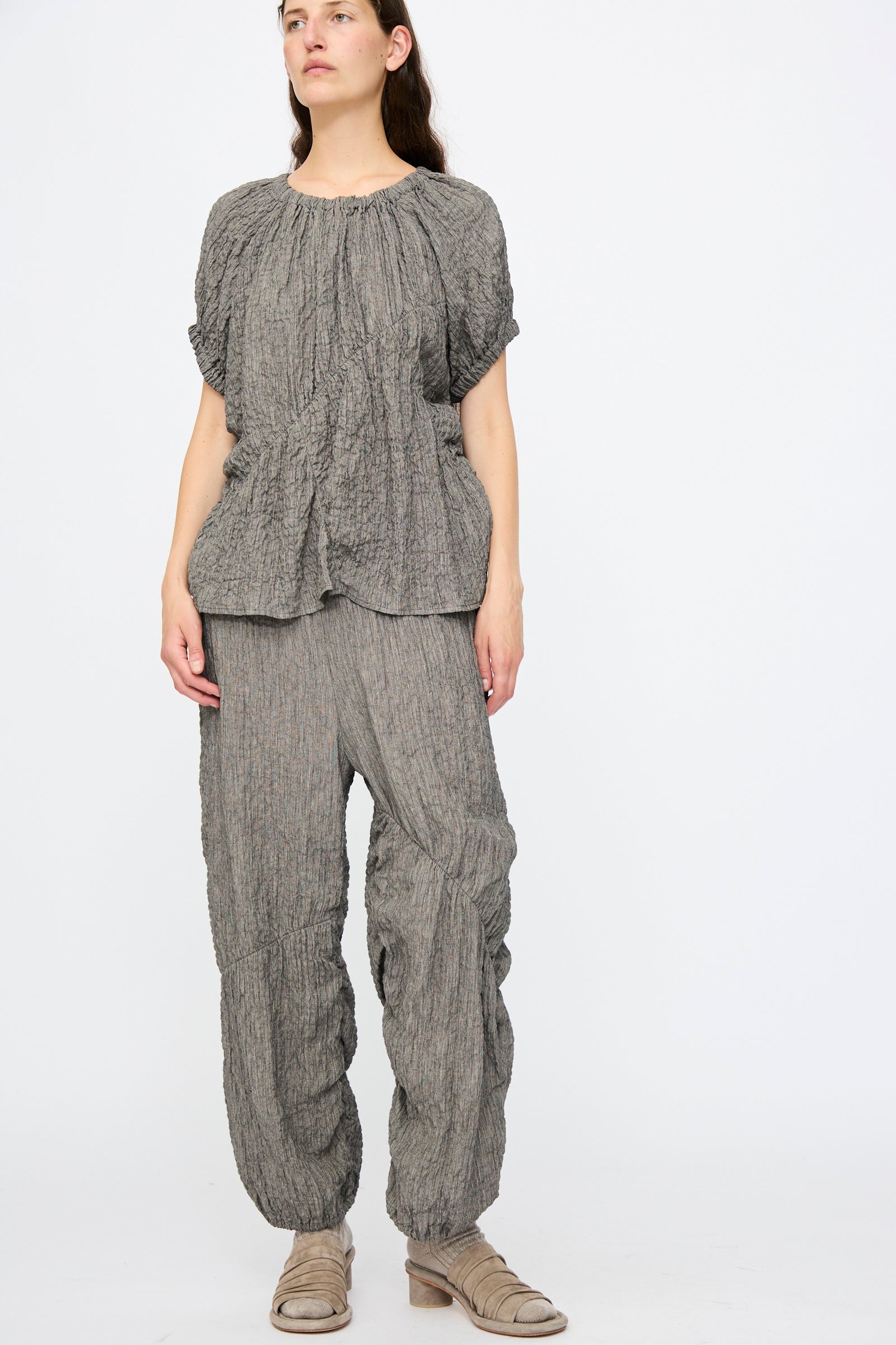 A person stands against a plain background, wearing Lauren Manoogian Shirred Pants in Charcoal, featuring a relaxed fit and elasticated waist, paired with a matching textured grey top and beige open-toe shoes.