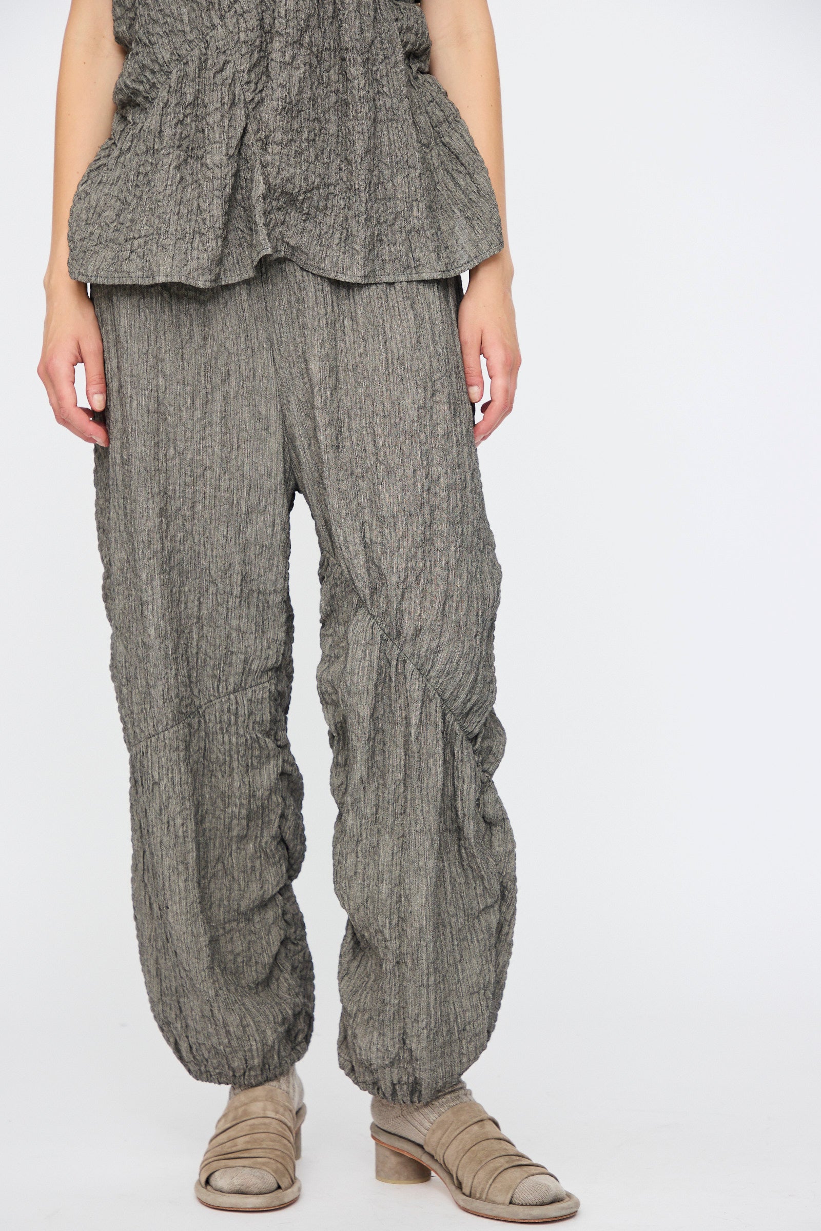 Model wears Lauren Manoogian’s Shirred Pants in Charcoal, a relaxed fit crinkled linen blend with elastic waist, paired with a matching top and beige ankle boots, standing before a plain white background.