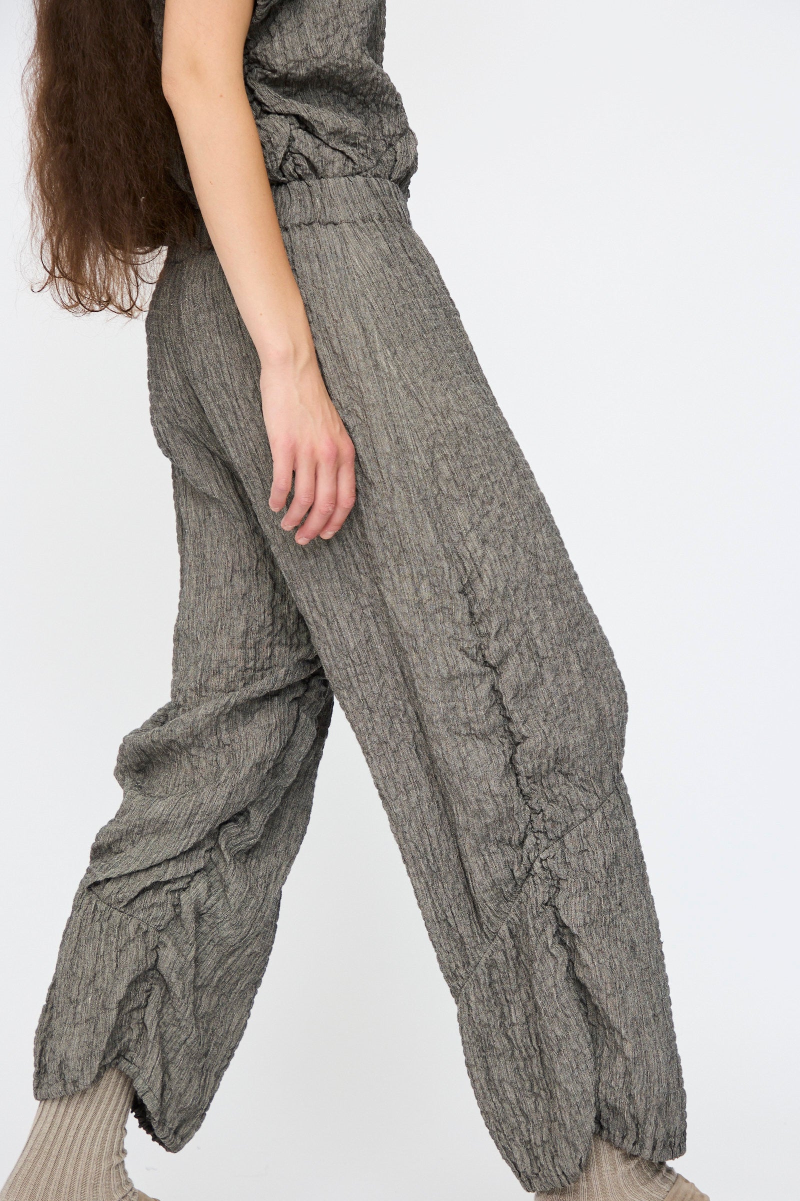 A person wears Lauren Manoogian's Shirred Pants in Charcoal, featuring a relaxed fit and elasticated waist, paired with light socks. Shown from mid-torso to feet against a plain background, their left arm rests by their side.