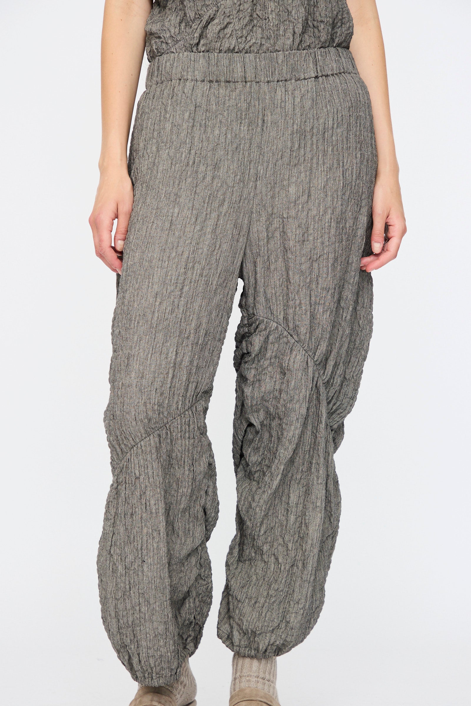 A person wearing Lauren Manoogian's Shirred Pants in Charcoal—relaxed fit with ruched detail and an elasticated waist—stands against a plain white background.