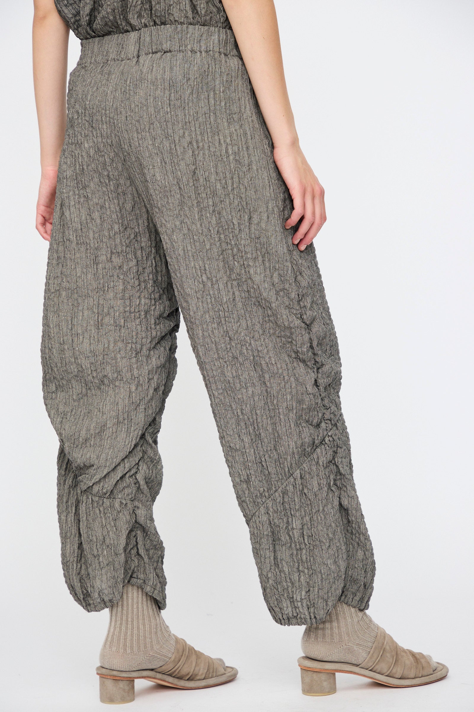 Model wearing Lauren Manoogian Shirred Pants in Charcoal—relaxed fit, crinkled linen blend with an elastic waist—paired with beige ribbed socks and open-toe heels, shown from the back against a plain backdrop.