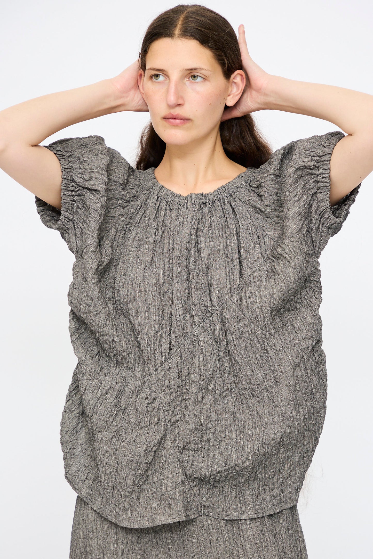 A woman models the Lauren Manoogian Shirred Top in Charcoal, featuring a loose, relaxed fit, textured linen blend, and gathered details, with her hands behind her head against a plain white background.